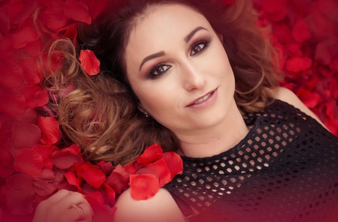 Portret kobiety z profesjonalnym makijażem, leżącej wśród czerwonych płatków róż. Uwagę przyciąga precyzyjny makijaż oczu i ust, podkreślający urodę modelki.