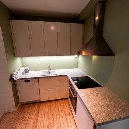 Solus Mateusz - Nowoczesna kuchnia z drewnianą podłogą, jasnymi szafkami i blatem w kolorze terazzo. Widoczny okap nad płytą indukcyjną i zlewozmywak z baterią. Ściany w odcieniu oliwkowej zieleni.