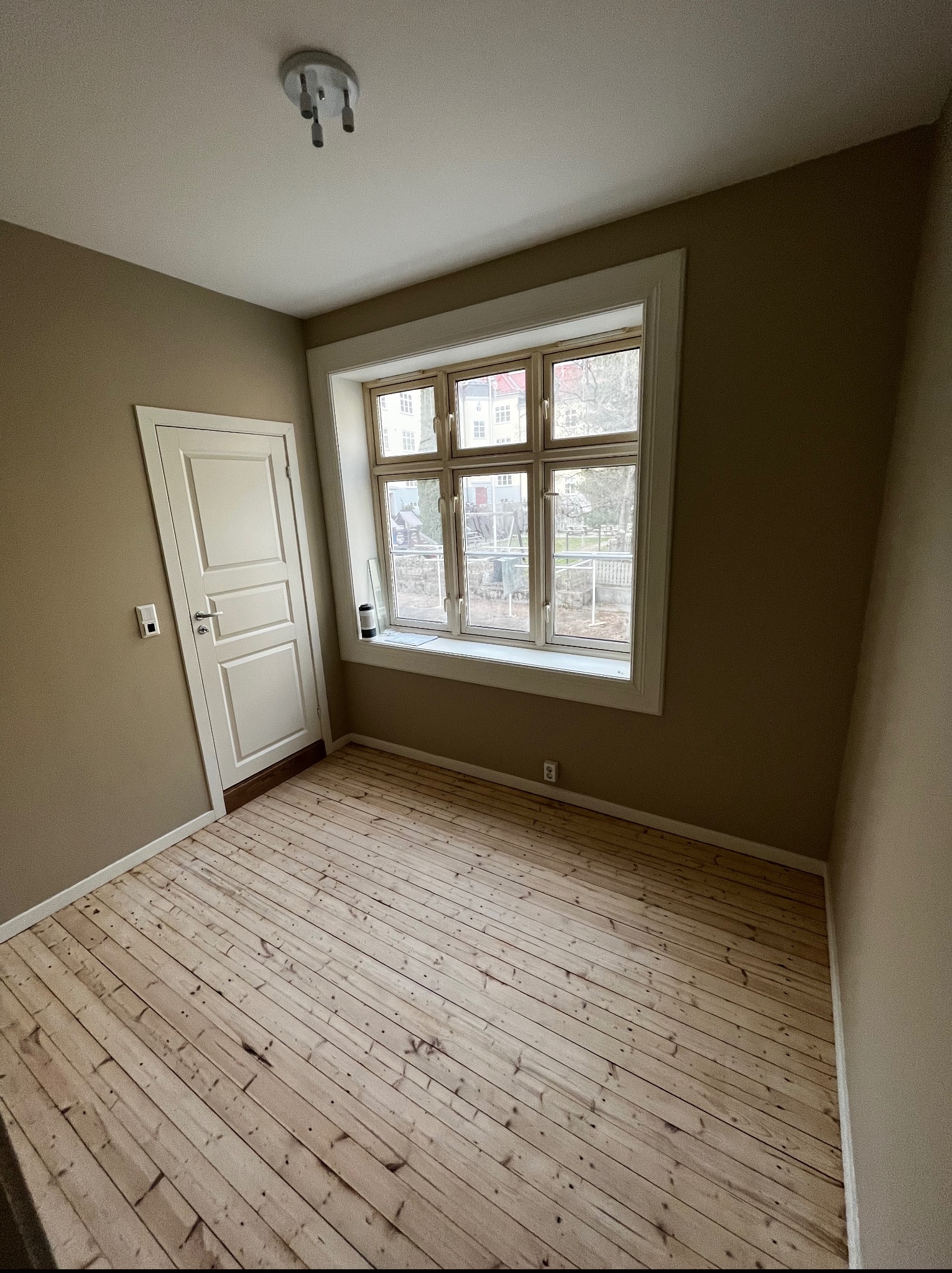 Pusty pokój z drewnianą podłogą i oknem z widokiem na zewnątrz. Ściany w odcieniach beżu, białe drzwi po lewej stronie. Minimalistyczne oświetlenie sufitowe.