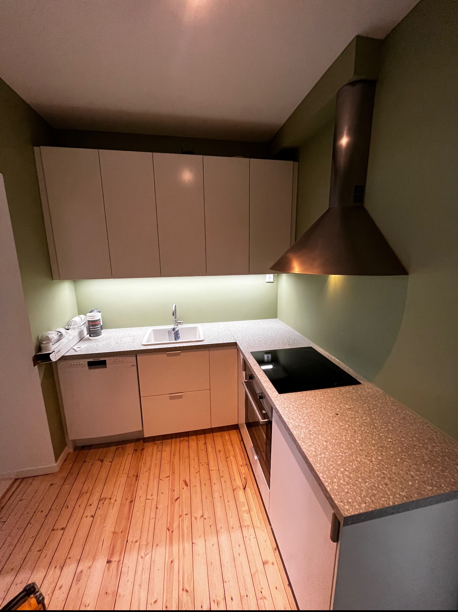 Nowoczesna kuchnia z drewnianą podłogą, jasnymi szafkami i blatem w kolorze terazzo. Widoczny okap nad płytą indukcyjną i zlewozmywak z baterią. Ściany w odcieniu oliwkowej zieleni.