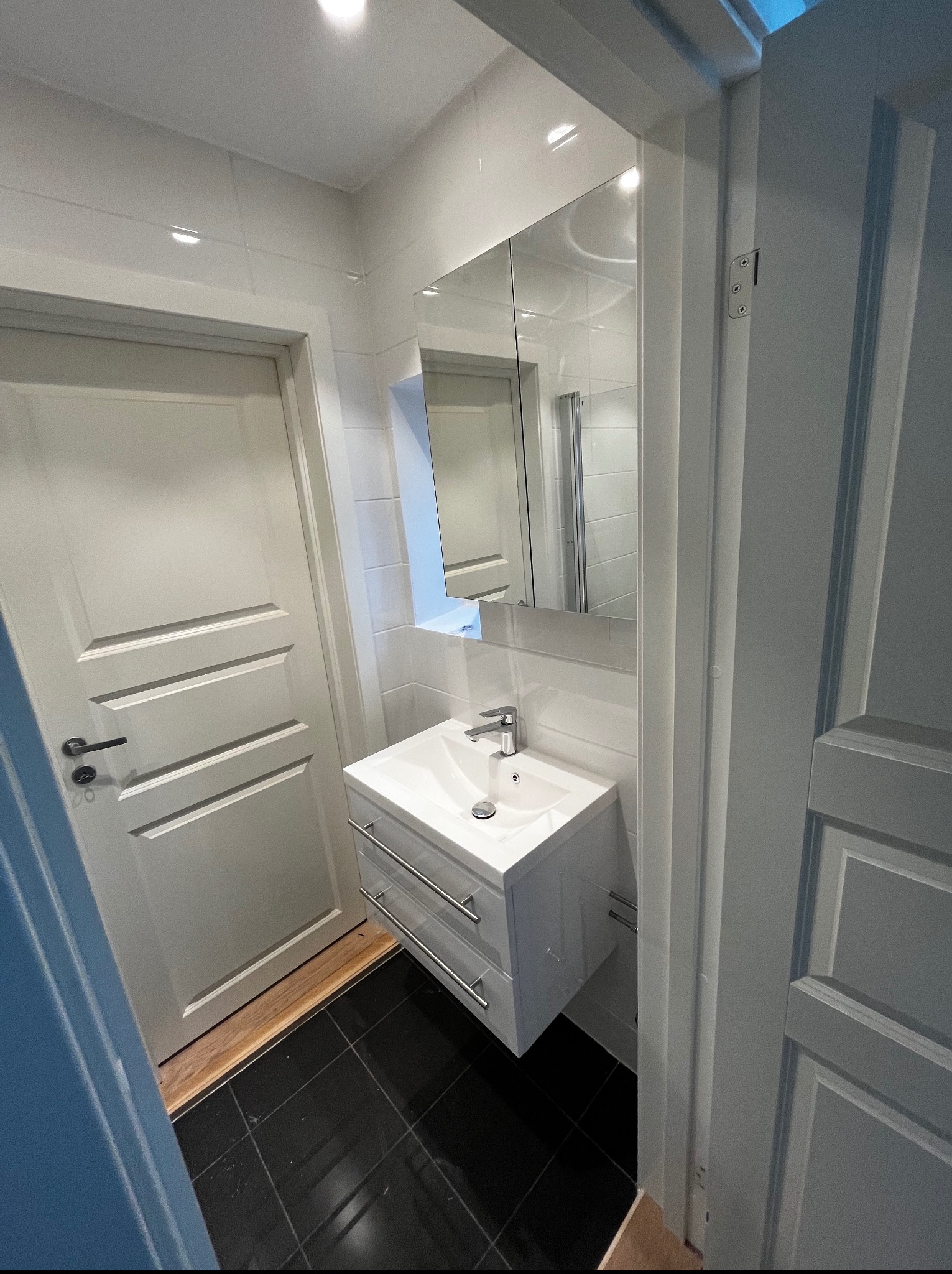 Biała umywalka z szafką w nowoczesnej łazience z czarną podłogą i lustrem. Widok na drzwi i fragment drugich drzwi. Minimalistyczny design.