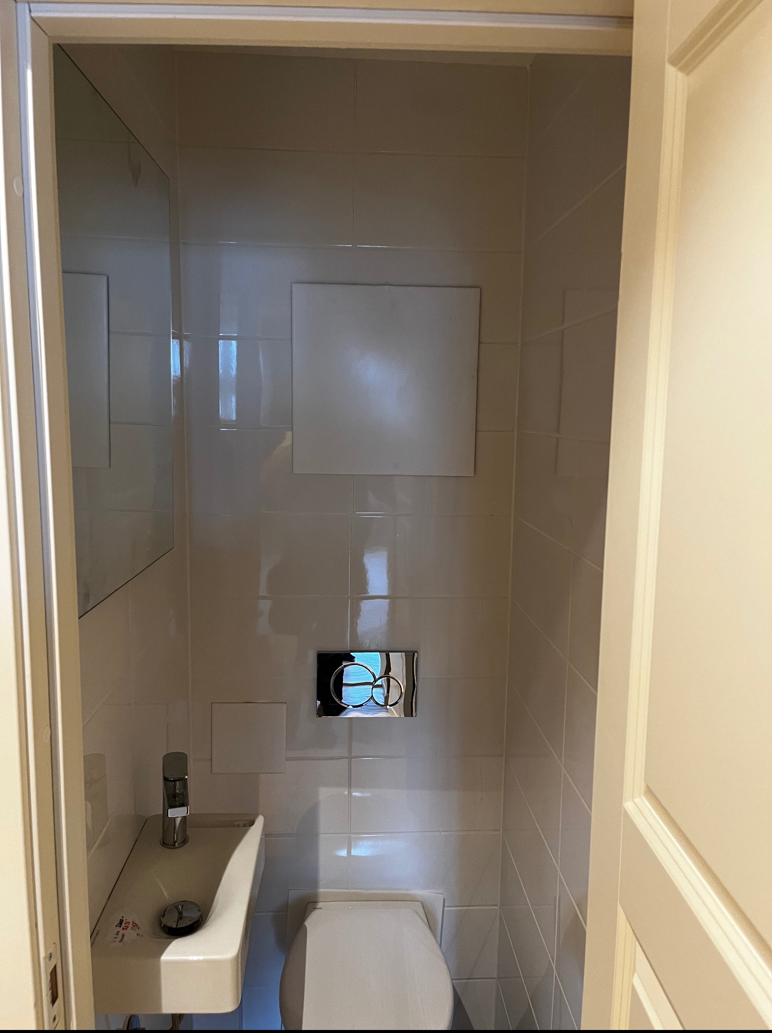 Mała, wykafelkowana toaleta z umywalką i lustrem. Widoczne białe płytki, toaleta i umywalka. Minimalistyczny design. Oświetlenie górne.