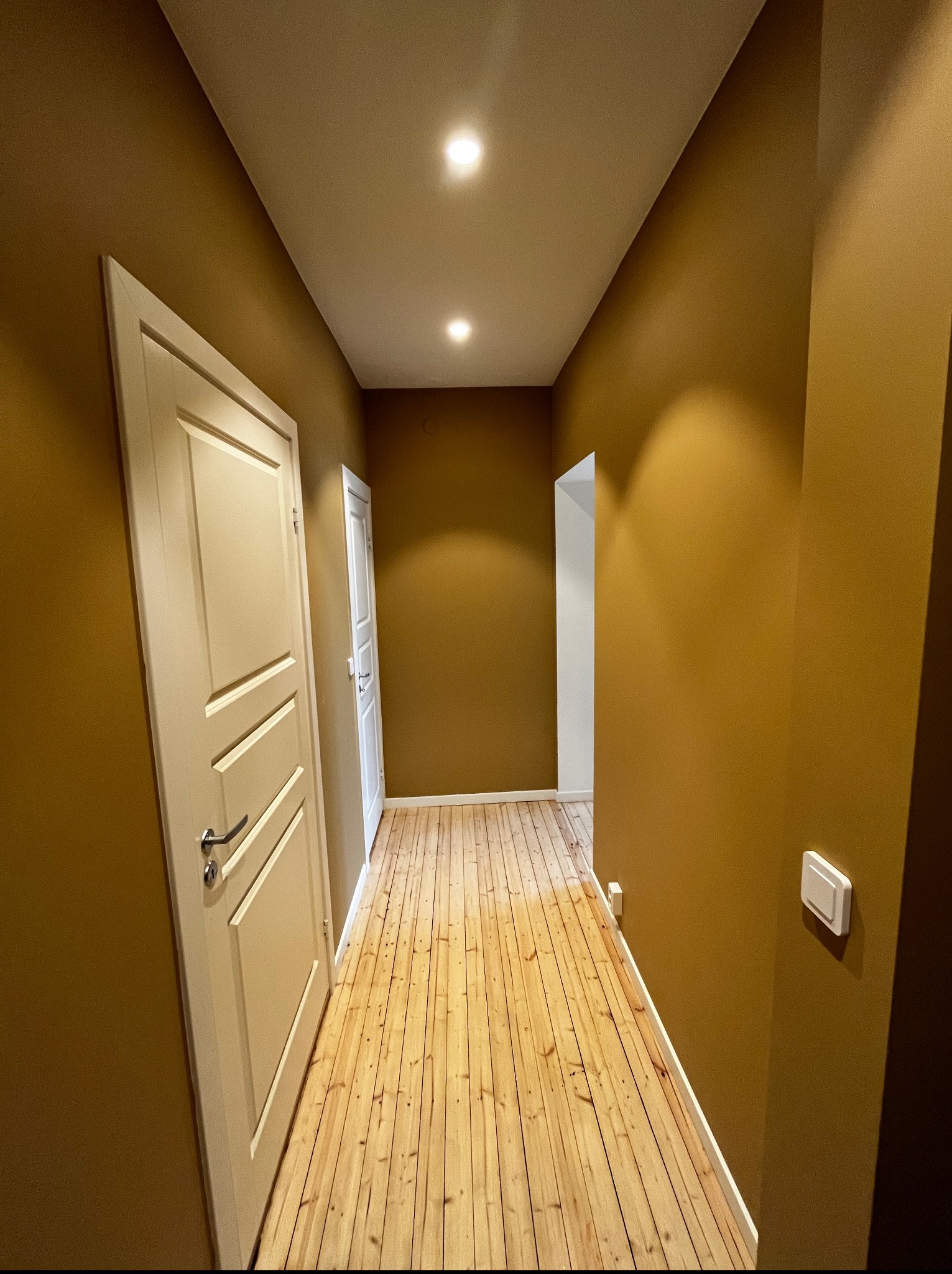 Korytarz z drewnianą podłogą i ścianami w odcieniach złota, oświetlony punktowo. Widoczne białe drzwi z ozdobnymi panelami i białe listwy przypodłogowe.