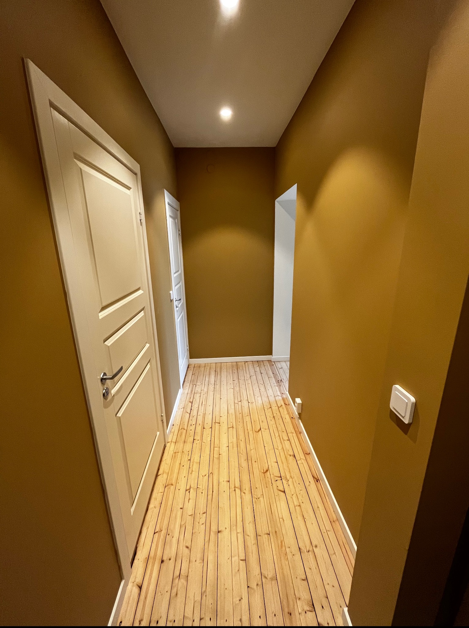 Korytarz z drewnianą podłogą i ścianami w odcieniach musztardowych. Widoczne białe drzwi z ozdobnymi panelami i białe wykończenia przy podłodze. Oświetlenie sufitowe.