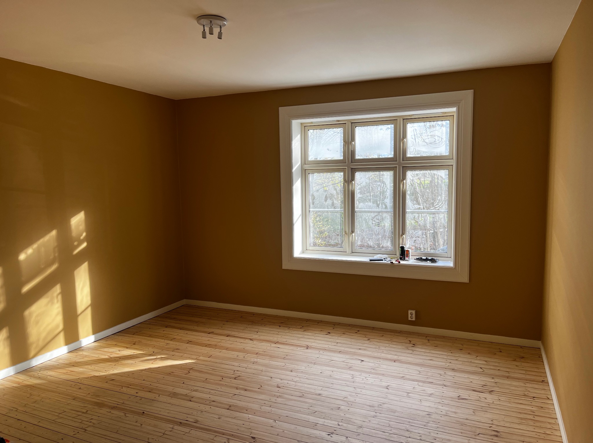 Pomalowane na musztardowy kolor wnętrze pokoju z drewnianą podłogą i oknem. Na parapecie widoczne narzędzia malarskie. Ściany gładkie, bez dekoracji. Promienie słońca na ścianie.