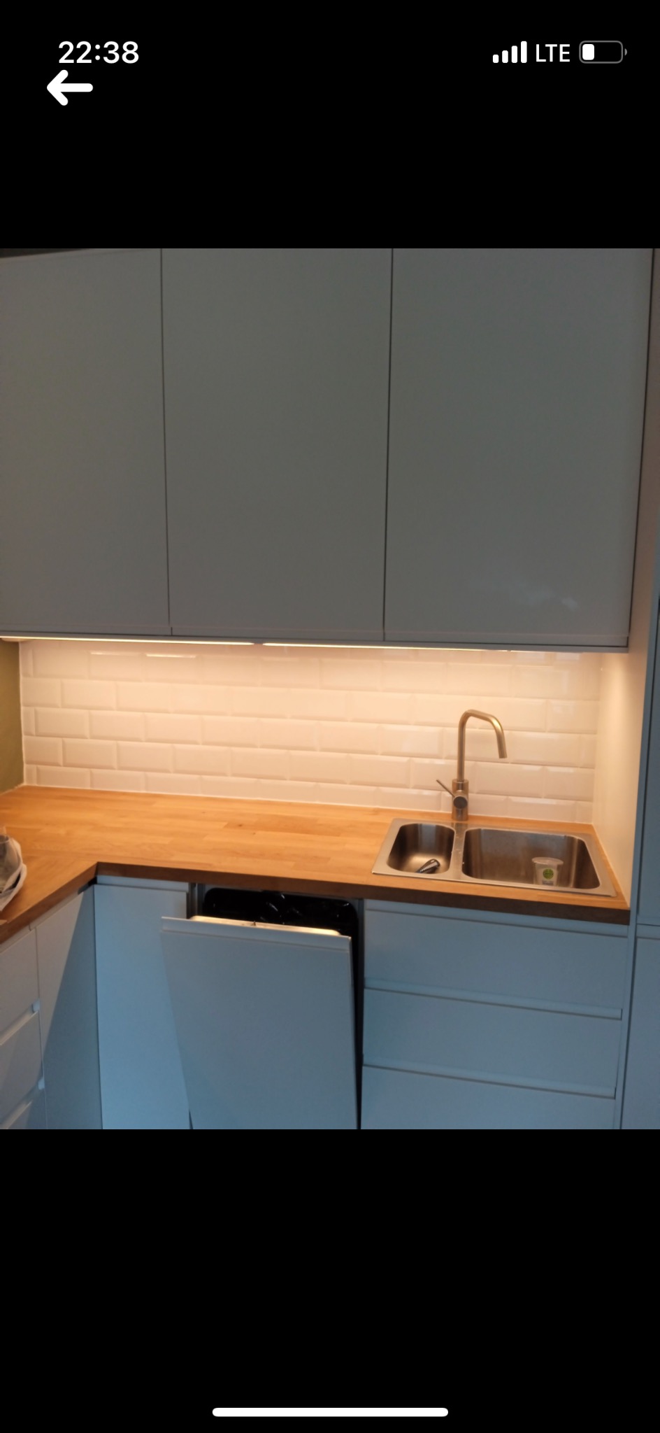 Nowoczesna kuchnia z białymi szafkami, drewnianym blatem i złotą baterią. Uwagę przyciągają białe, prostokątne płytki na ścianie i oświetlenie podszafkowe.