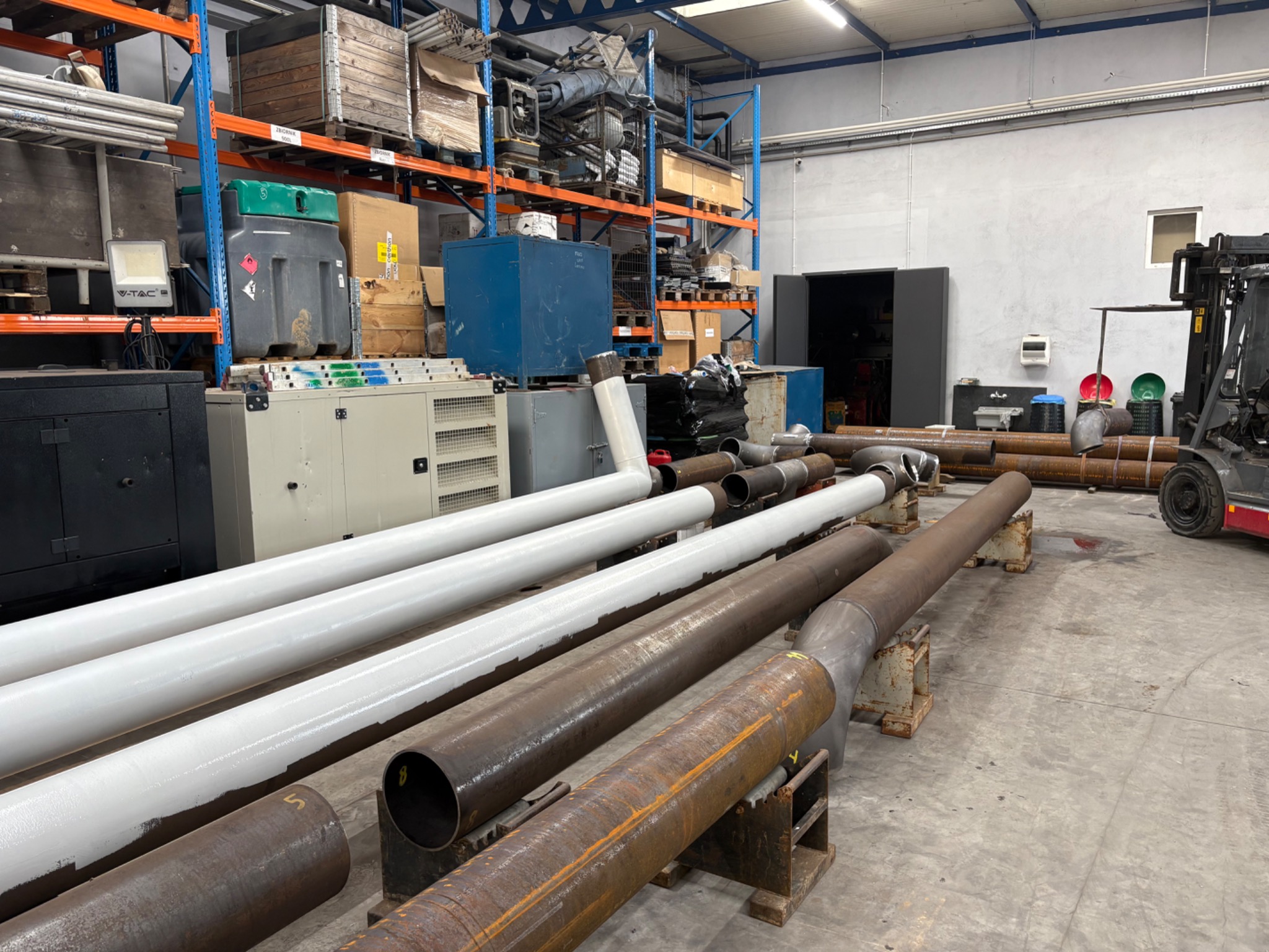 Rury stalowe, niektóre pomalowane na biało, leżące na metalowych podporach w hali produkcyjnej. W tle regały z materiałami i wózki widłowe.