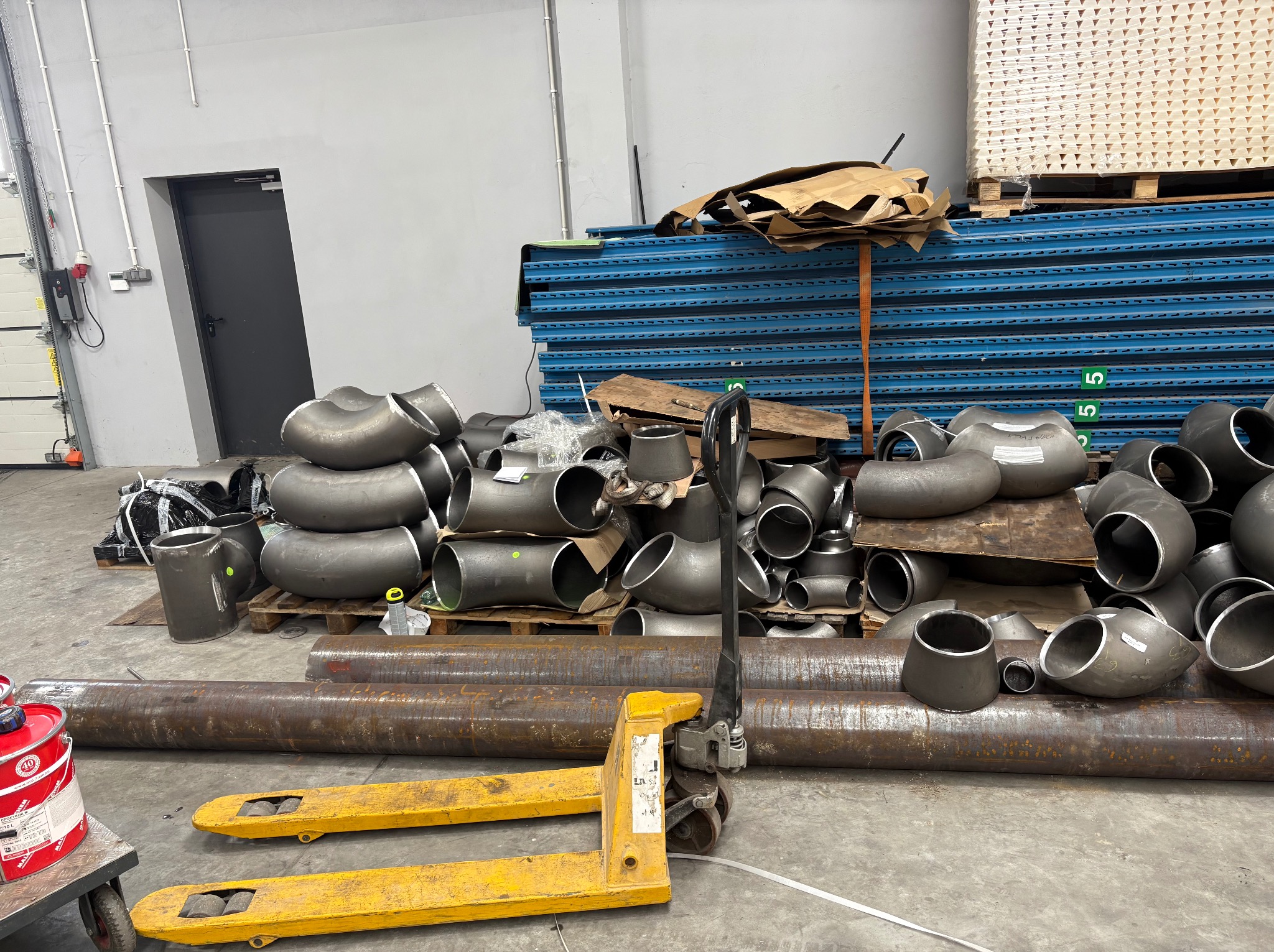 Stalowe kolanka i rury na palecie, żółty wózek paletowy, magazynowe tło z regałami i drzwiami. Elementy instalacji przemysłowej przed montażem.