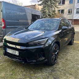 Mateusz Jankowski - Błyszczący, czarny samochód Cupra Formentor zaparkowany na trawie przed budynkiem. Widoczny szary van w tle i tablica rejestracyjna PY 1309J.