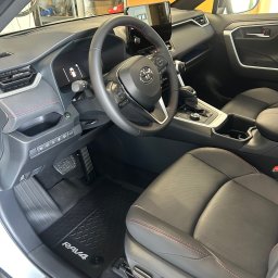 Mateusz Jankowski - Wnętrze Toyoty RAV4 Plug-in Hybrid: czarna skórzana tapicerka z czerwonymi przeszyciami, gumowe dywaniki RAV4, widok na deskę rozdzielczą i kierownicę.