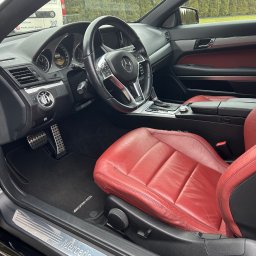 Mateusz Jankowski - Wnętrze czarnego Mercedesa z czerwoną skórzaną tapicerką fotela kierowcy, widoczne detale deski rozdzielczej i pedałów. Na zewnątrz zaparkowany biały bus.