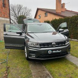 Mateusz Jankowski - Szary Volkswagen Tiguan z otwartymi drzwiami i bagażnikiem, zaparkowany na trawniku przed domem. Widoczny wąż ogrodowy i logo firmy detailingowej na tablicy rejestracyjnej.