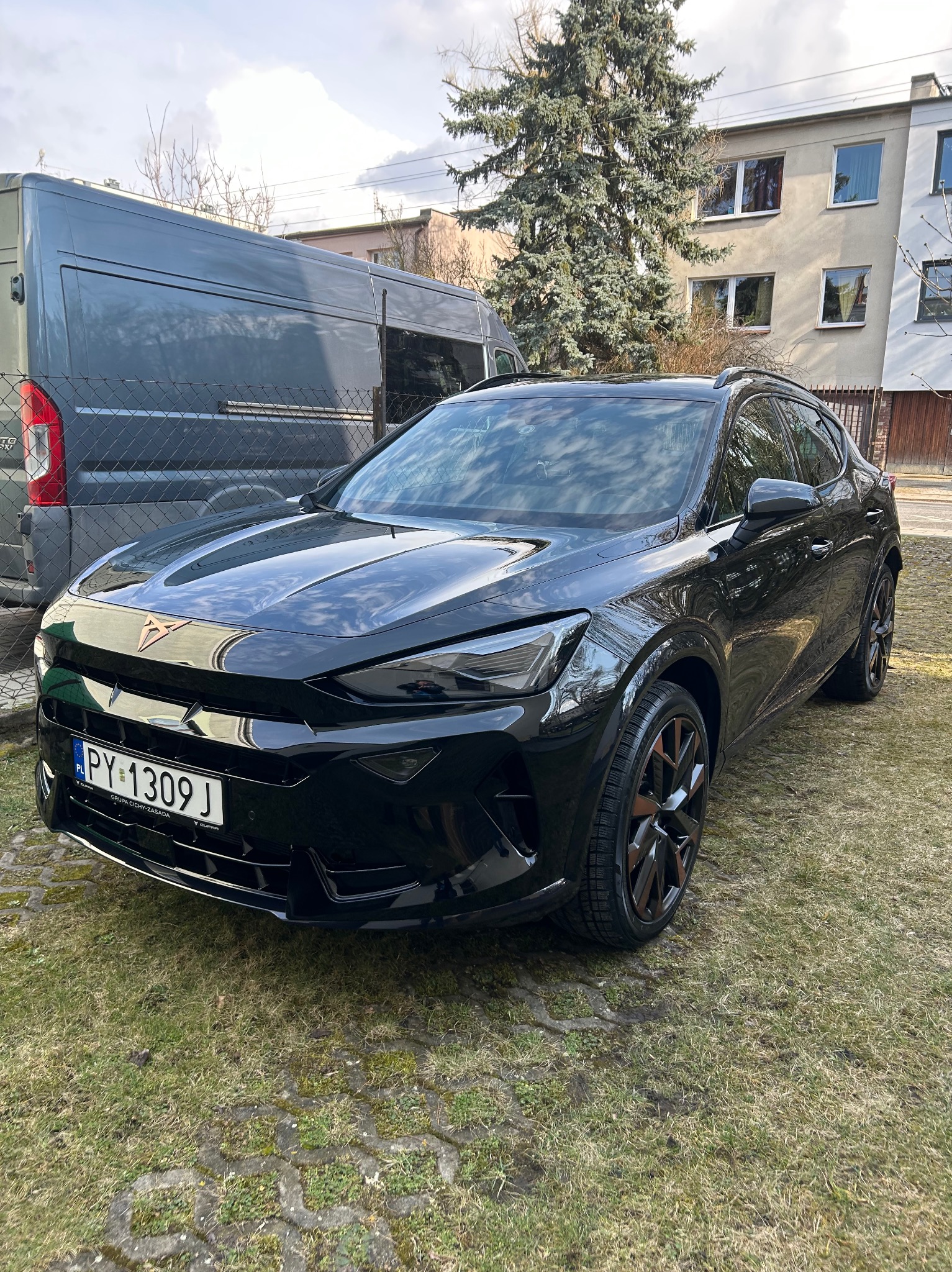 Błyszczący, czarny samochód Cupra Formentor zaparkowany na trawie przed budynkiem. Widoczny szary van w tle i tablica rejestracyjna PY 1309J.