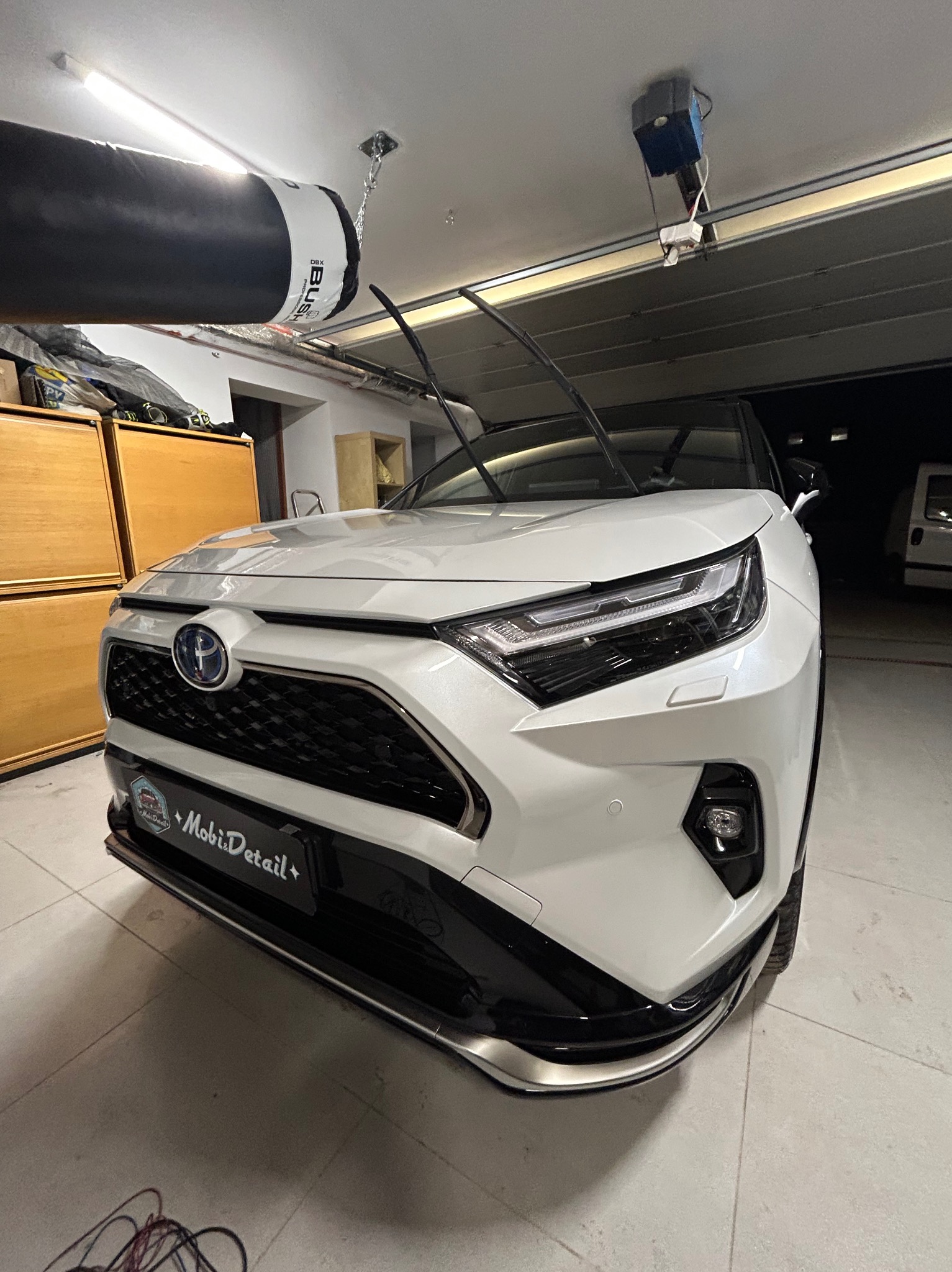 Biały SUV Toyota RAV4 po myciu, zaparkowany w garażu z podniesioną bramą. Widoczne detale karoserii i reflektory. Na przednim zderzaku naklejka MobiDetail.