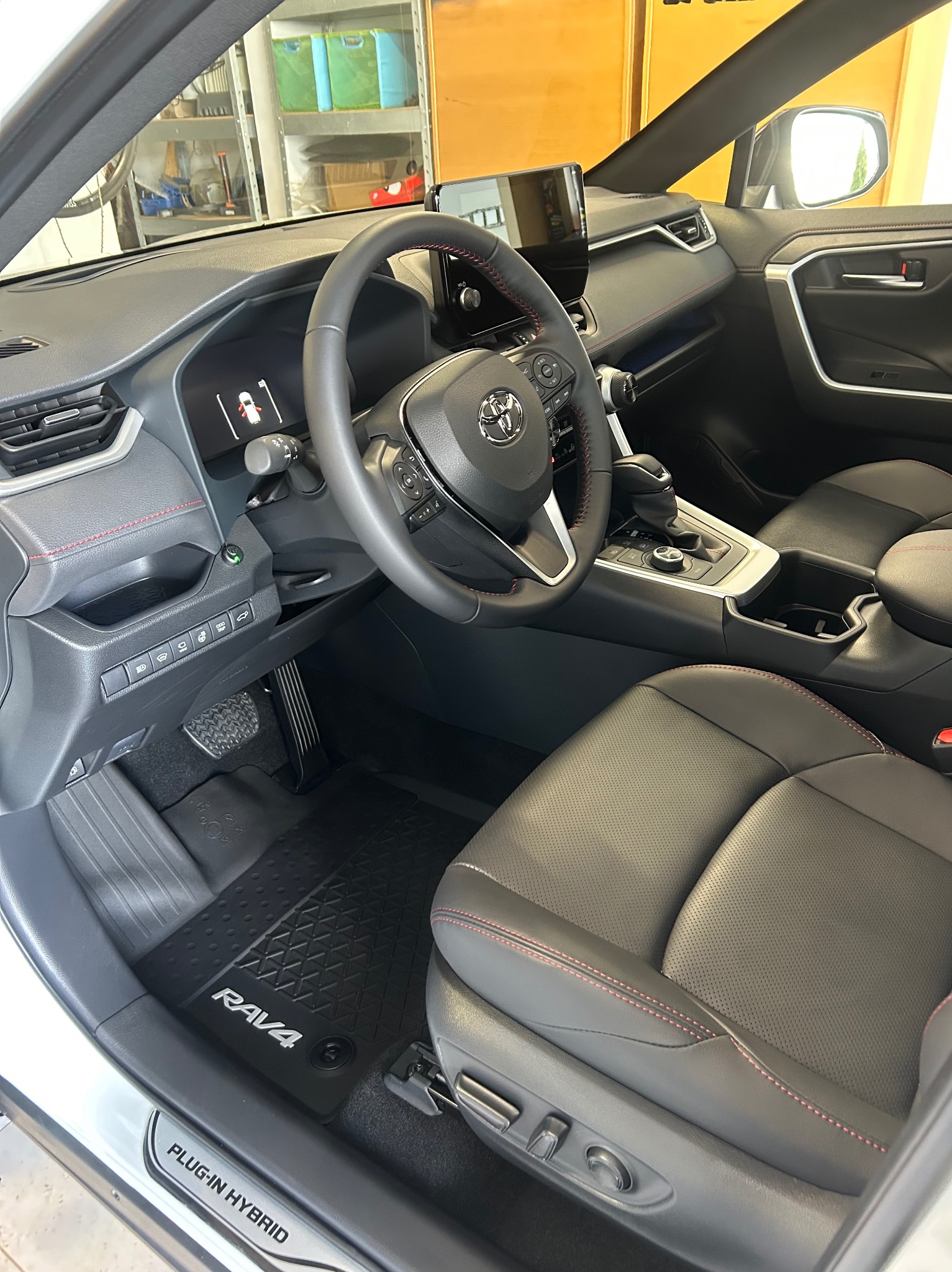 Wnętrze Toyoty RAV4 Plug-in Hybrid: czarna skórzana tapicerka z czerwonymi przeszyciami, gumowe dywaniki RAV4, widok na deskę rozdzielczą i kierownicę.