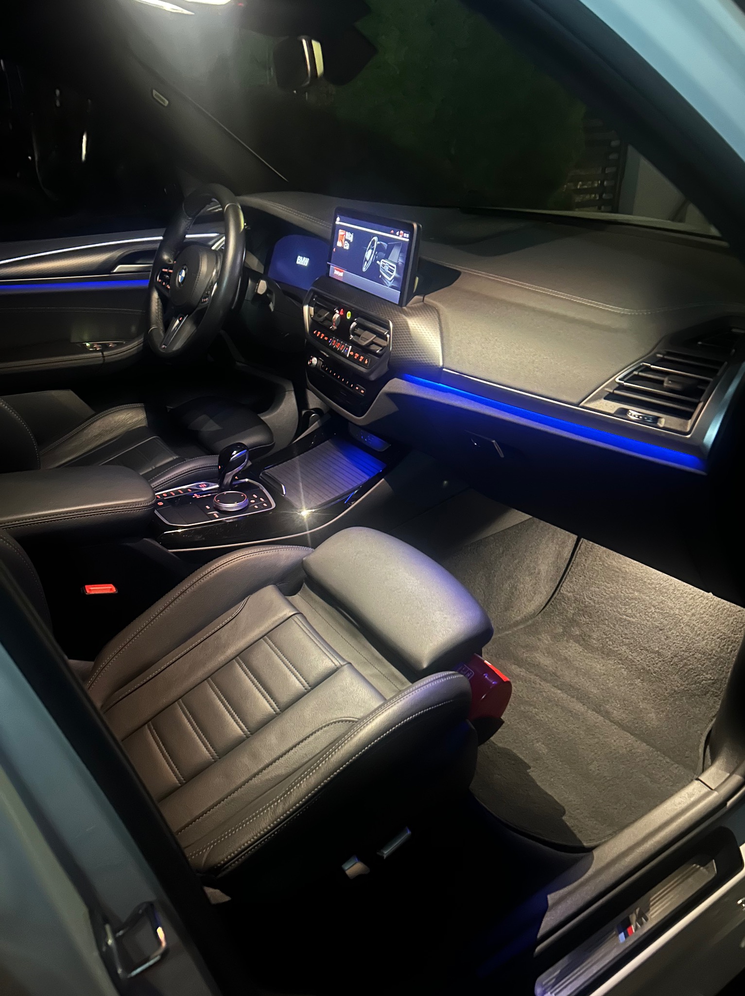 Wnętrze luksusowego samochodu BMW z czarną skórzaną tapicerką i subtelnym, niebieskim podświetleniem deski rozdzielczej i konsoli środkowej. Widoczne fotele i kokpit.