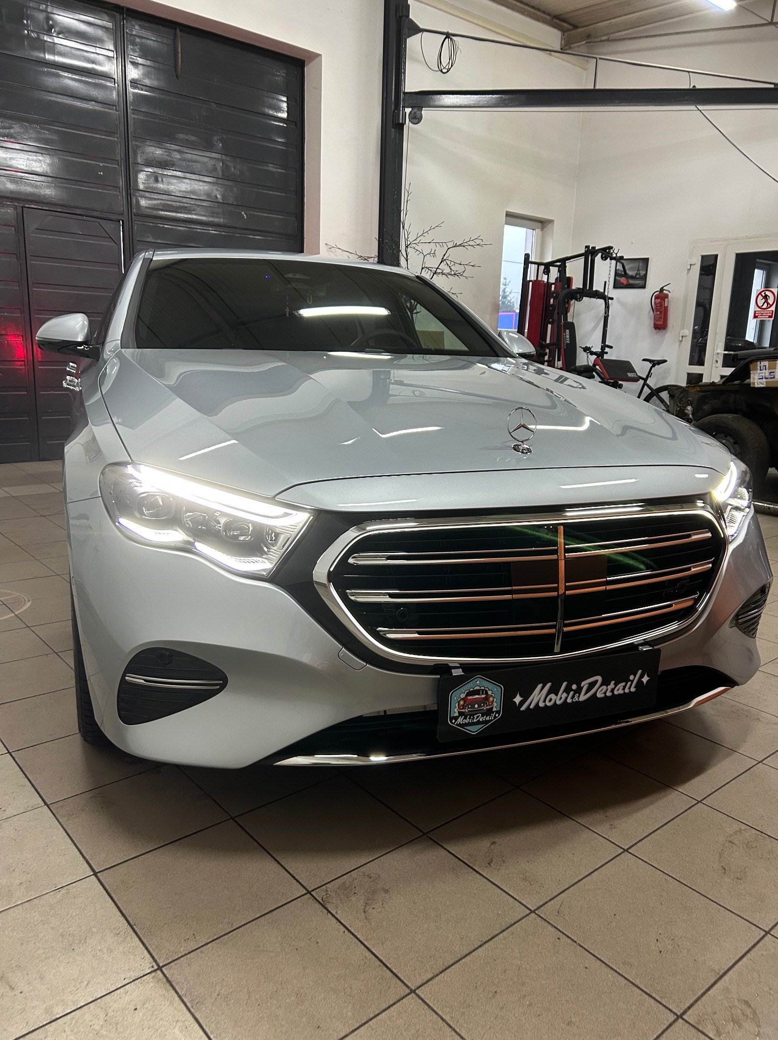 Błyszczący, srebrny Mercedes po detailingu, z widocznym logo firmy na tablicy rejestracyjnej, na tle garażu z wyposażeniem do ćwiczeń.