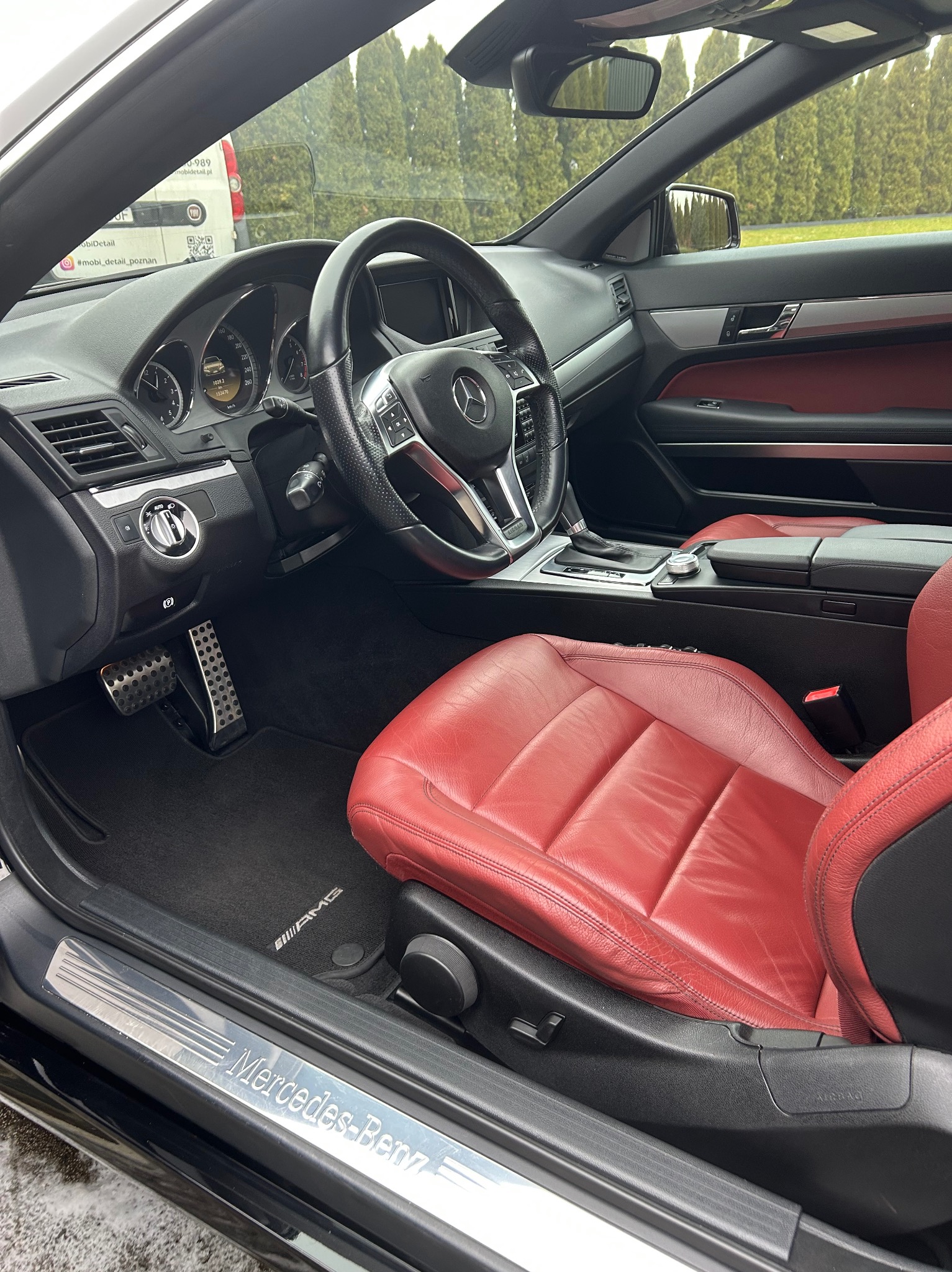 Wnętrze czarnego Mercedesa z czerwoną skórzaną tapicerką fotela kierowcy, widoczne detale deski rozdzielczej i pedałów. Na zewnątrz zaparkowany biały bus.
