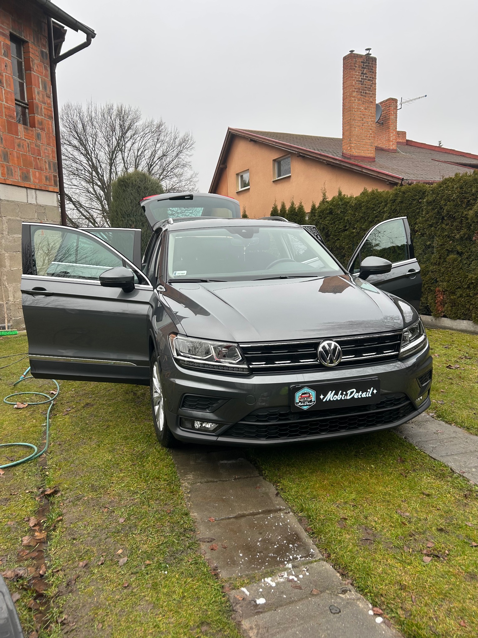 Szary Volkswagen Tiguan z otwartymi drzwiami i bagażnikiem, zaparkowany na trawniku przed domem. Widoczny wąż ogrodowy i logo firmy detailingowej na tablicy rejestracyjnej.