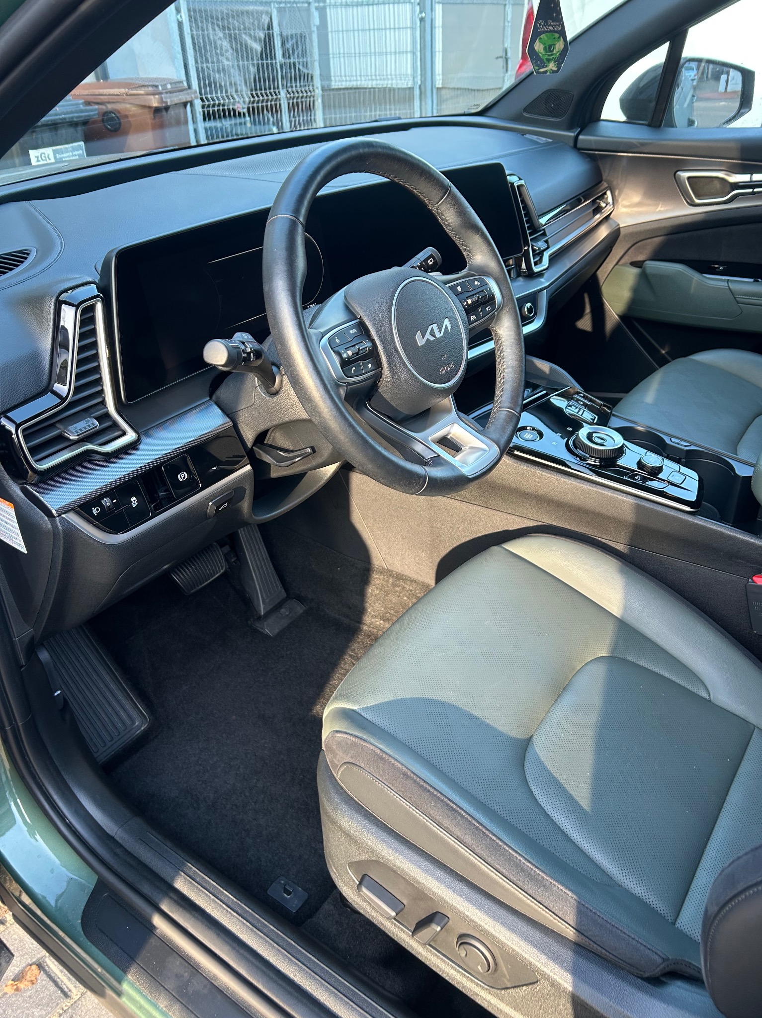 Wnętrze nowoczesnego samochodu Kia Sportage: kierownica z logo, skórzane fotele w odcieniach szarości, deska rozdzielcza z ekranem dotykowym i elementami sterowania.