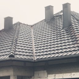 Paw-Bud - Szary dach z dachówki ceramicznej z trzema kominami wentylacyjnymi, widok z dołu na budynek w trakcie budowy, z widocznymi bloczkami betonowymi.