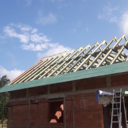 Paw-Bud - Budowa dachu z drewnianą konstrukcją i zielonym pokryciem na murowanym budynku w trakcie wznoszenia, widoczne rusztowania i drabina.