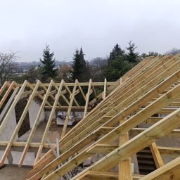 Paw-Bud - Konstrukcja drewniana dachu w trakcie budowy, widok z góry na krokwie i murłatę, w tle zieleń drzew i zabudowa miejska w pochmurny dzień.