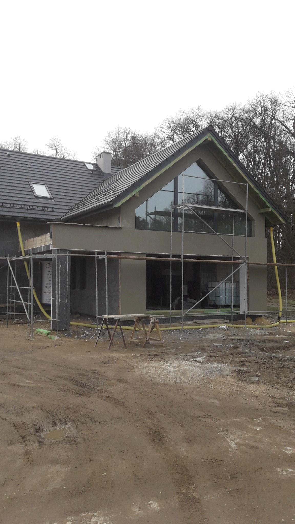 Surowy dom z dużym oknem trójkątnym w szczycie, w trakcie prac elewacyjnych. Widoczne rusztowania i przygotowane ściany do dalszych etapów wykończenia.