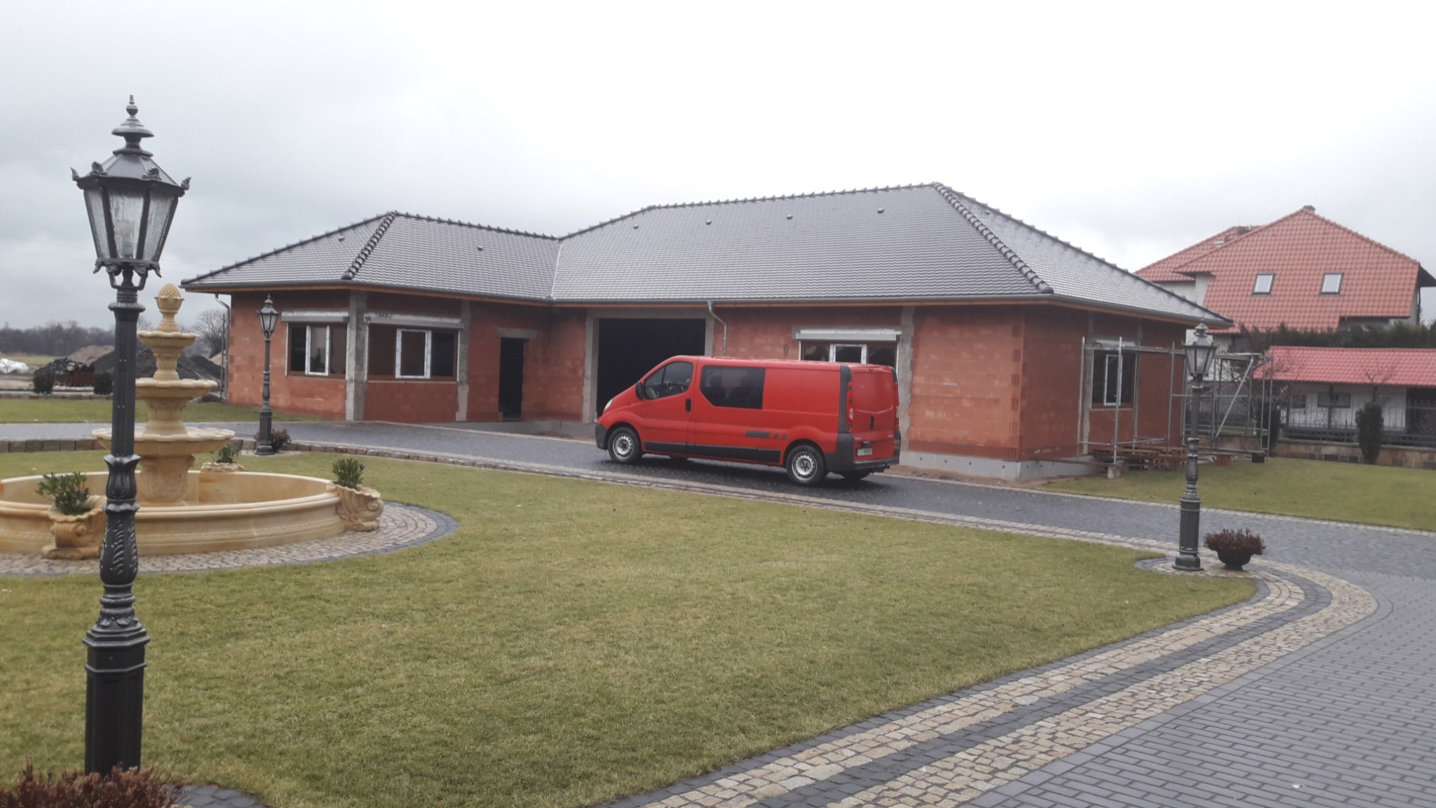 Niedokończony dom z czerwonej cegły z szarym dachem, czerwony van na podjeździe, ozdobna fontanna i latarnia, trawnik. Dom w trakcie budowy.