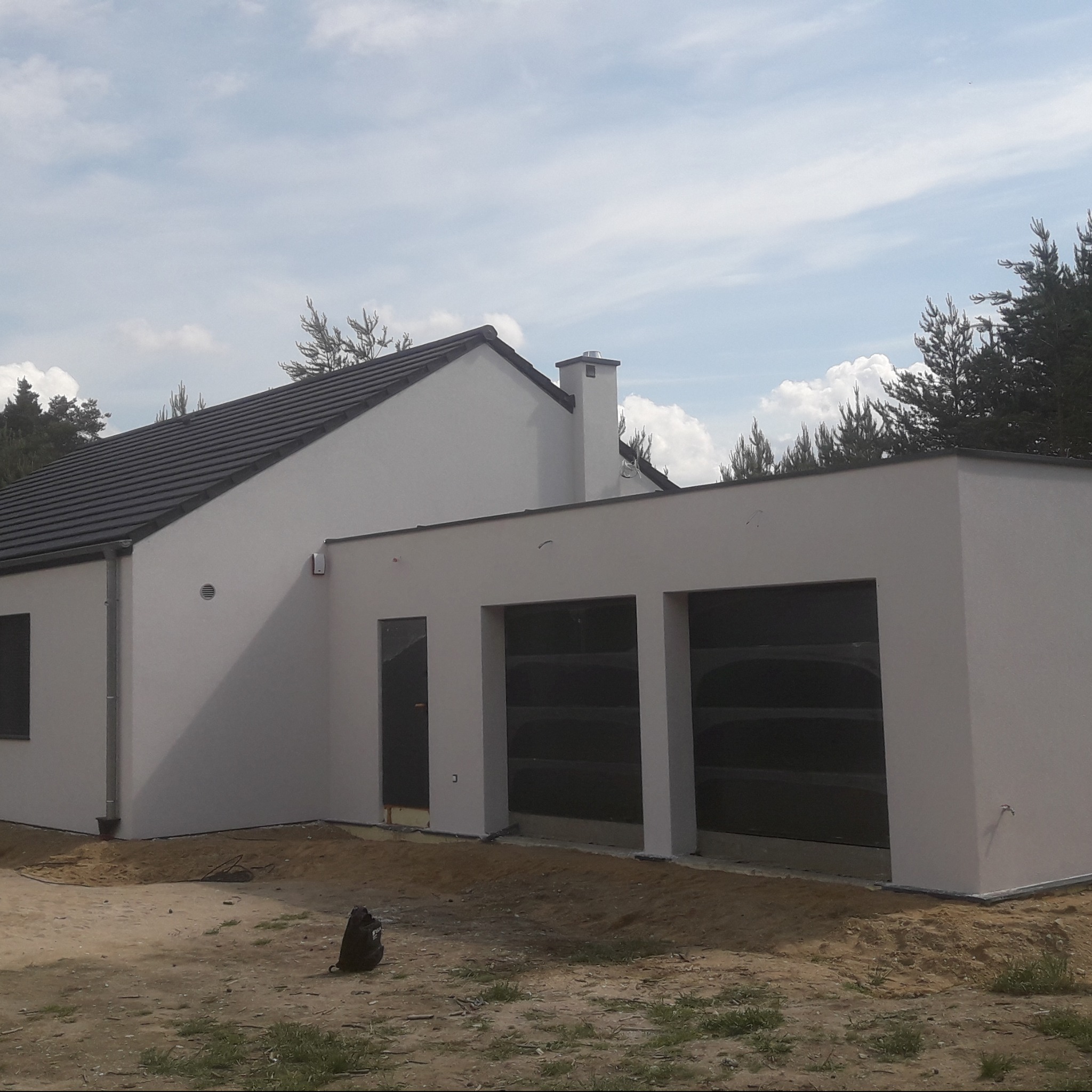 Nowoczesny dom z białymi ścianami i ciemnym dachem w słoneczny dzień. Widoczne duże okna garażowe i komin. Otoczenie w trakcie prac wykończeniowych.