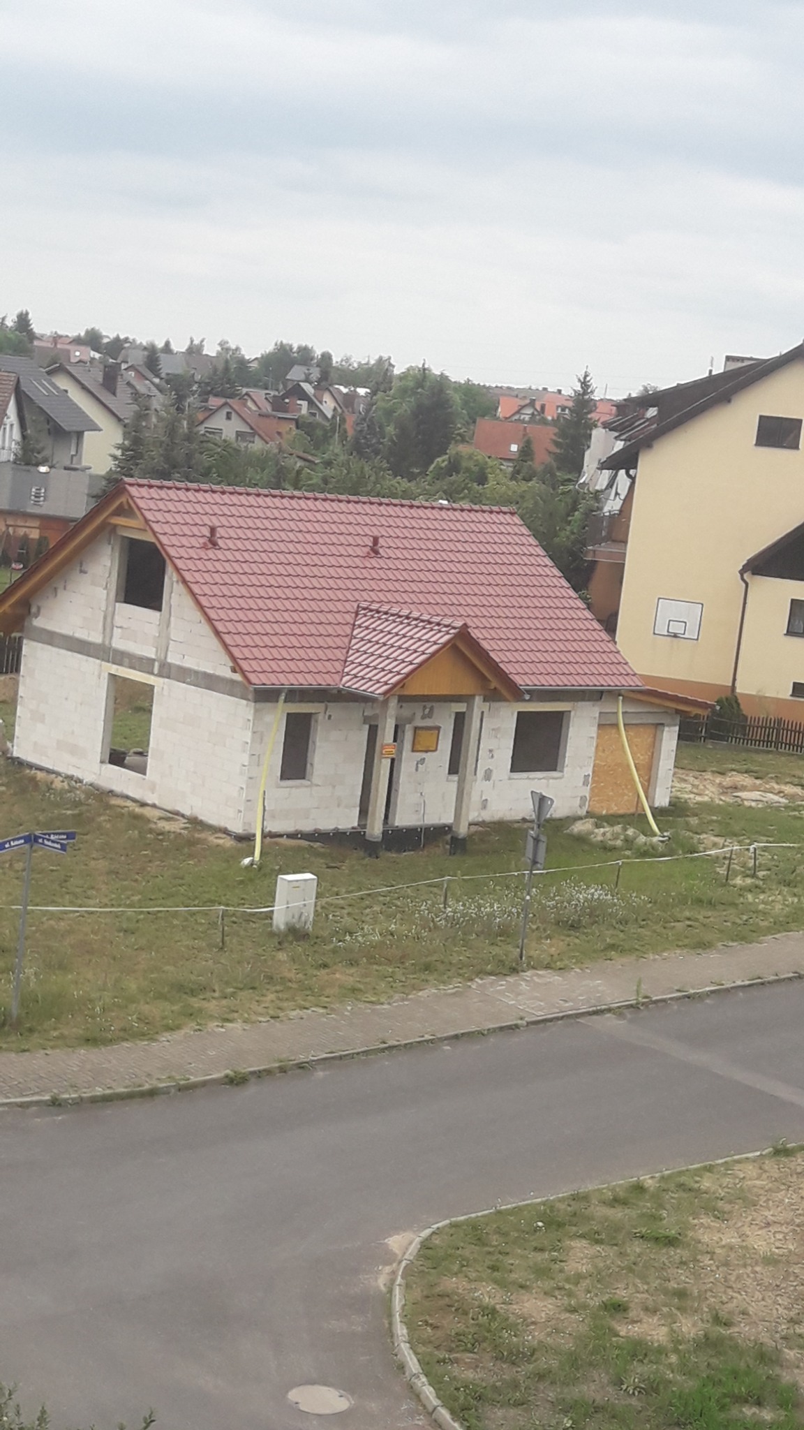 Niedokończony dom z białego pustaka z czerwonym dachem w otoczeniu innych budynków i zieleni, widok z góry.