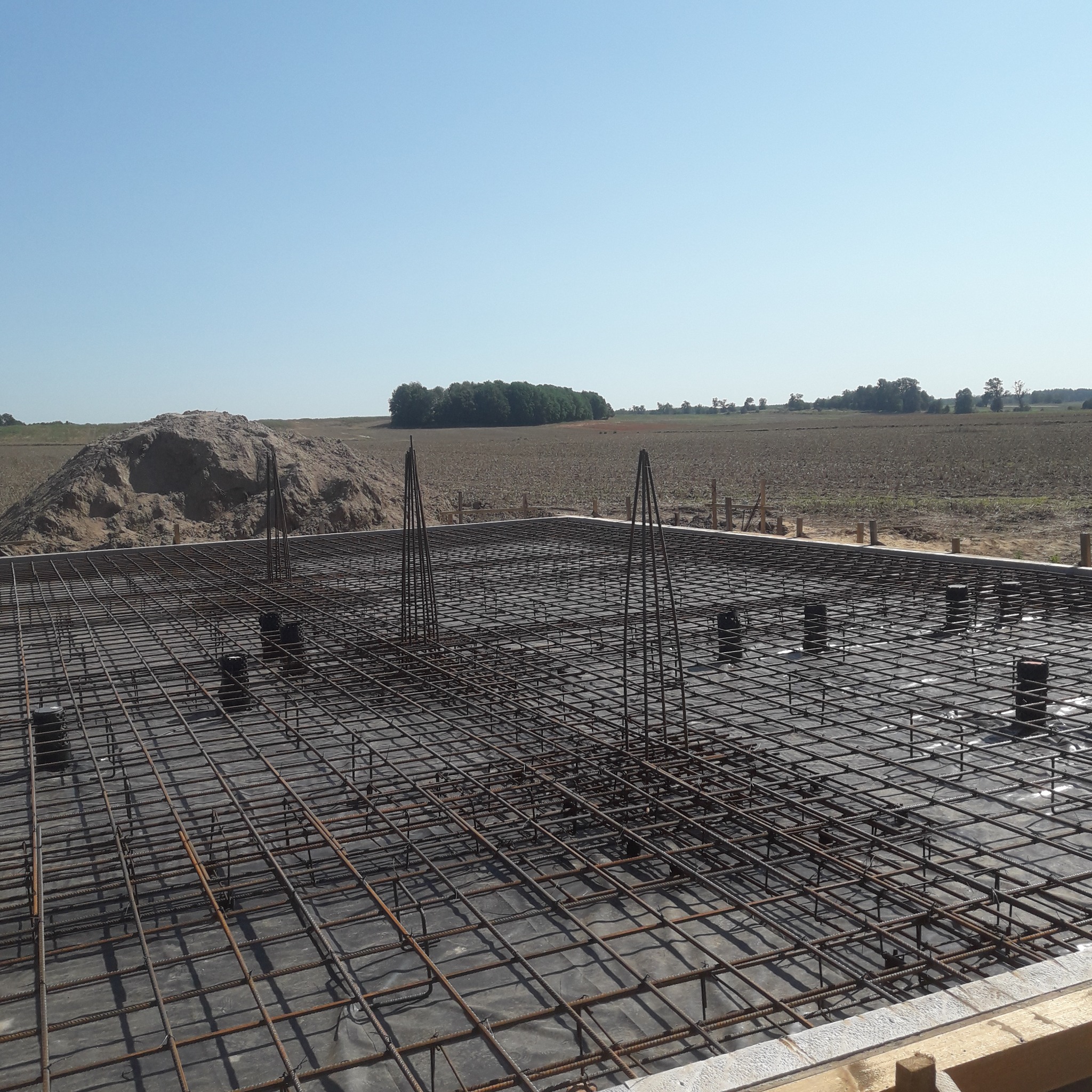 Zbrojenie fundamentów domu jednorodzinnego z widoczną siatką stalową, pionowymi prętami i elementami dystansowymi na tle pola i błękitnego nieba.