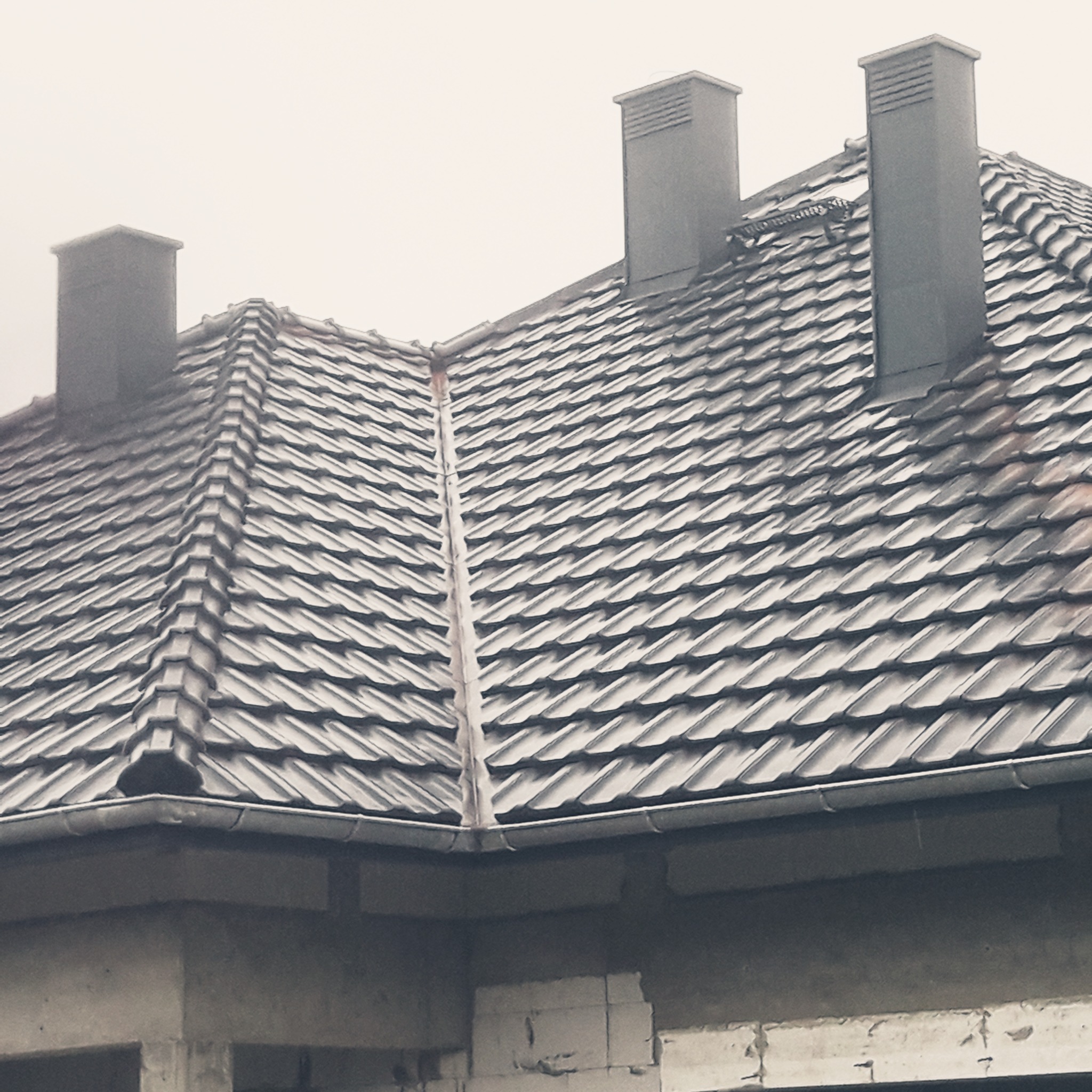 Szary dach z dachówki ceramicznej z trzema kominami wentylacyjnymi, widok z dołu na budynek w trakcie budowy, z widocznymi bloczkami betonowymi.