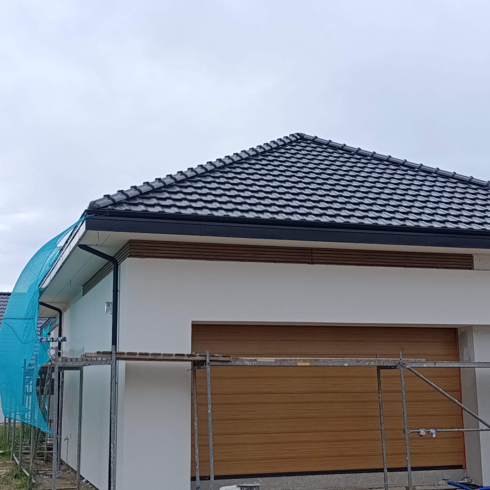 Dom jednorodzinny z ciemną dachówką, białą elewacją i drewnianą bramą garażową. Widoczne rusztowania i siatka ochronna, sugerujące prace wykończeniowe.