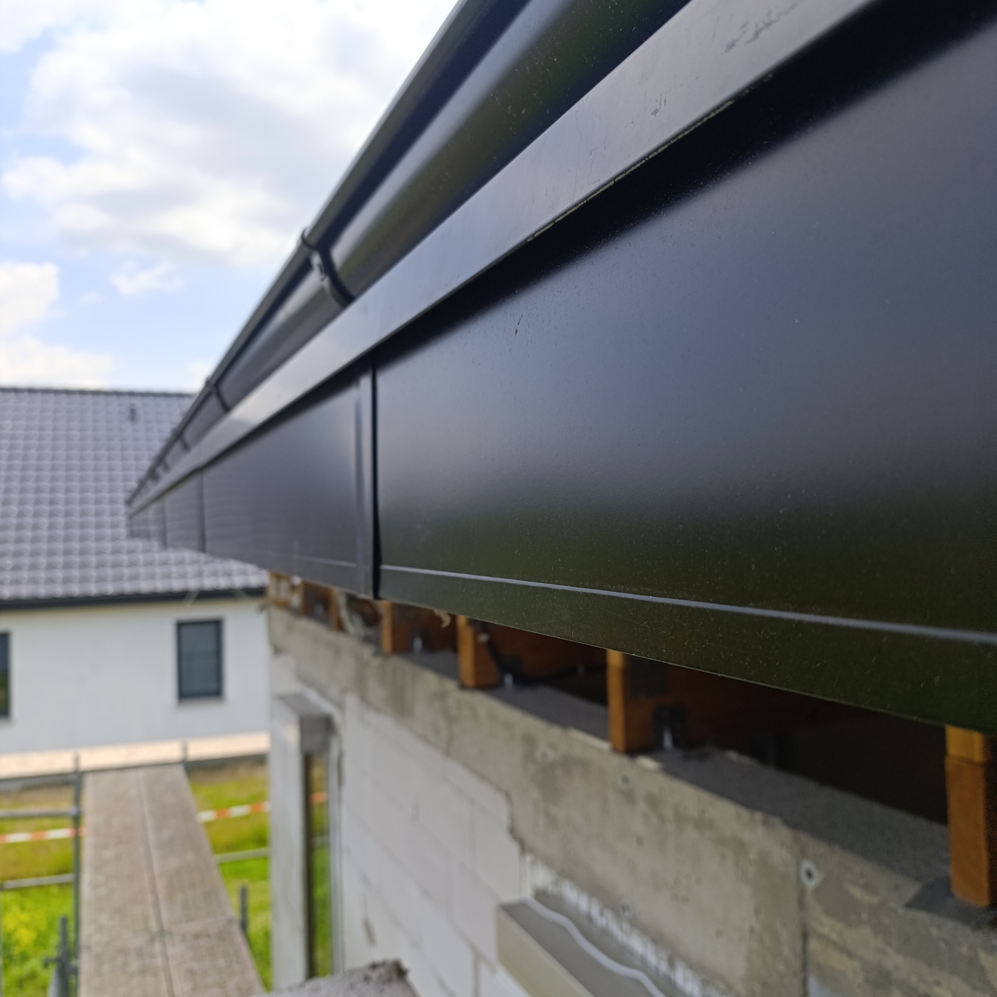 Czarny system rynnowy zamontowany na budynku w trakcie budowy, widoczny fragment rusztowania i fragment dachu sąsiedniego domu. Ujęcie z dołu, pochmurne niebo.