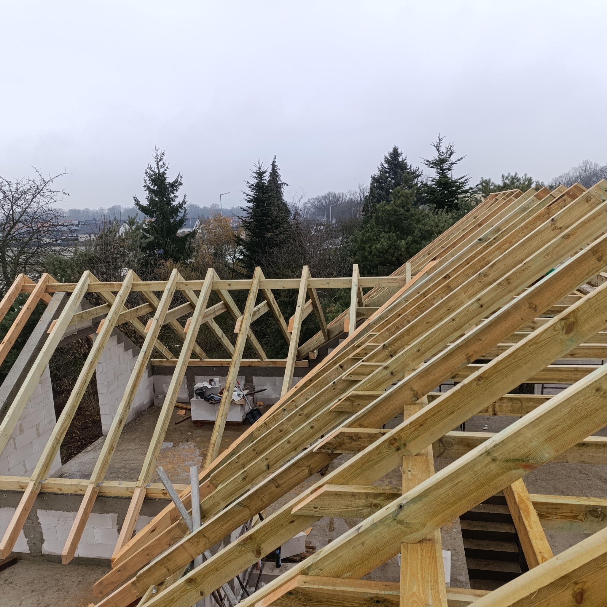 Konstrukcja drewniana dachu w trakcie budowy, widok z góry na krokwie i murłatę, w tle zieleń drzew i zabudowa miejska w pochmurny dzień.
