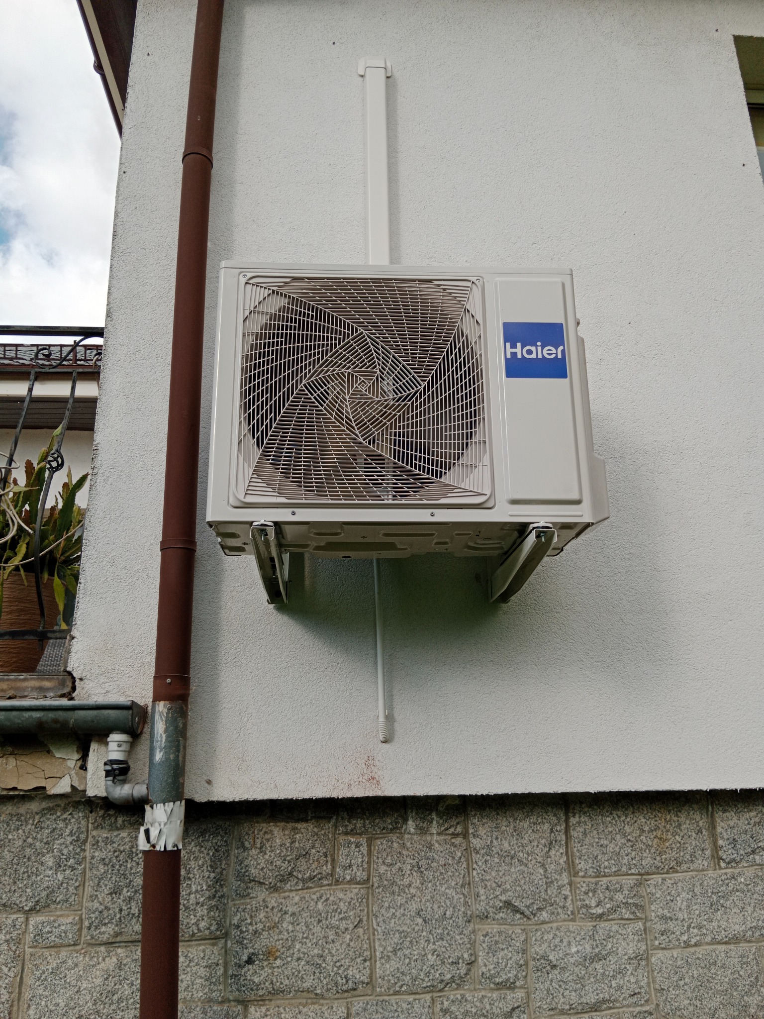 Biała jednostka zewnętrzna klimatyzacji Haier zamontowana na ścianie budynku z kamienną podmurówką i brązową rurą spustową. Widoczne mocowania i fragment rośliny w tle.
