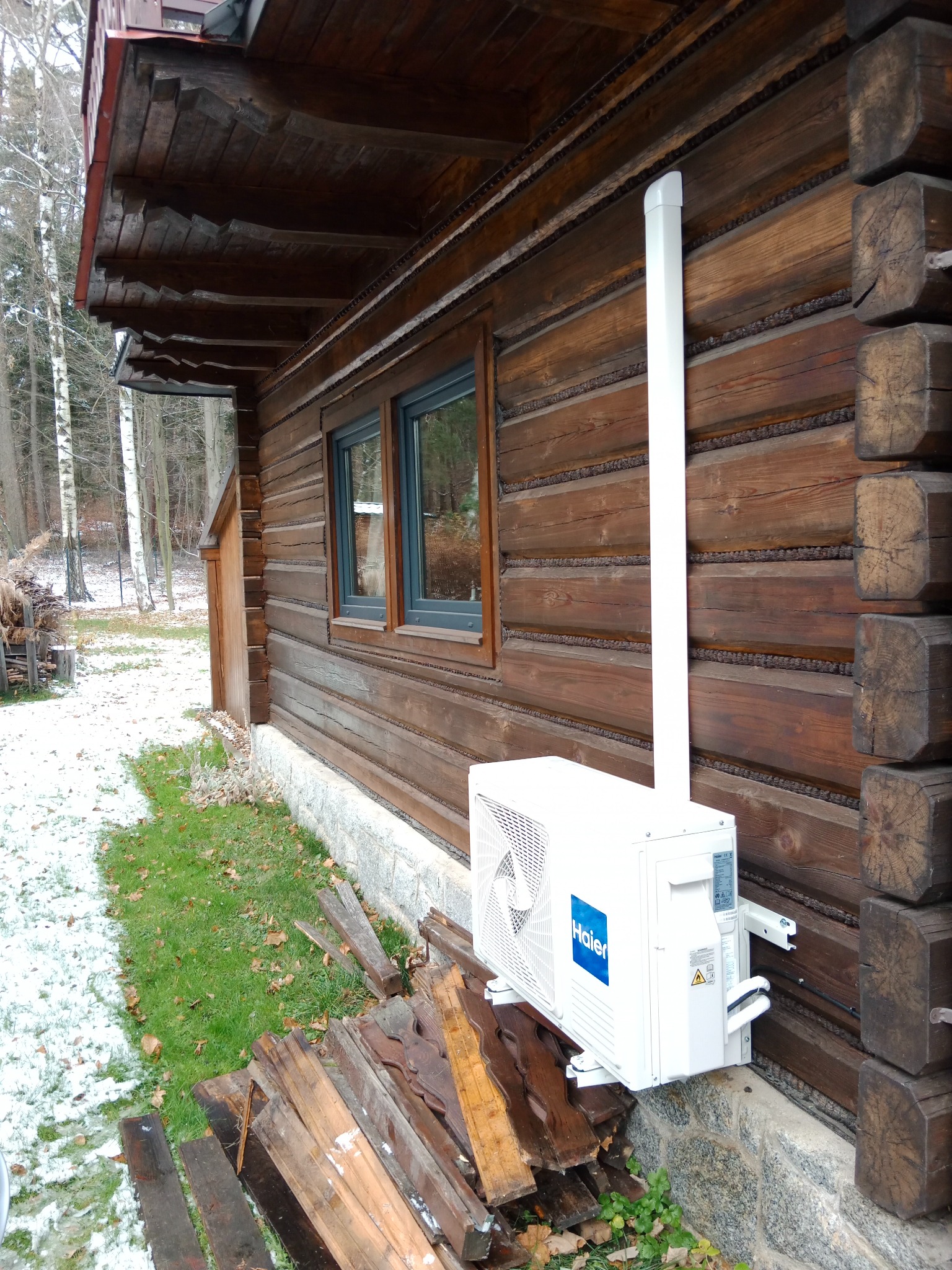 Biała jednostka zewnętrzna klimatyzacji Haier zamontowana na drewnianej ścianie domu z bali, z widoczną instalacją odprowadzającą. Częściowo ośnieżony teren.