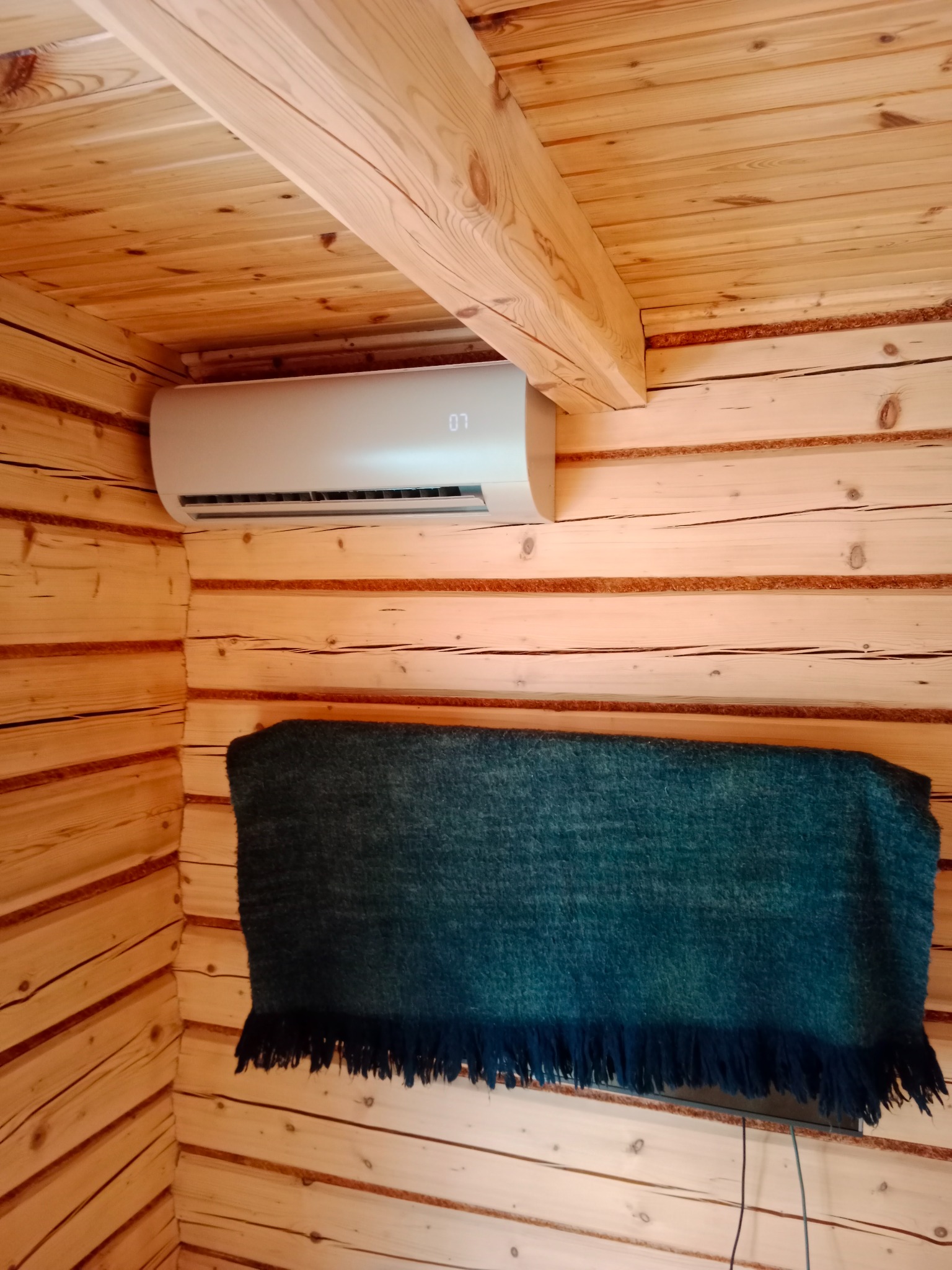 Biała klimatyzacja ścienna w drewnianym wnętrzu, widoczna część ściany z bali i fragment ciemnego koca z frędzlami. Minimalistyczny design w rustykalnym otoczeniu.