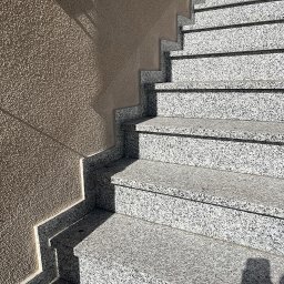 Kamieniarstwo RADEK - Zewnętrzne schody z jasnego, nakrapianego kamienia prowadzą w górę, przylegając do ściany o szorstkiej fakturze. Metalowa poręcz widoczna w lewym górnym rogu.