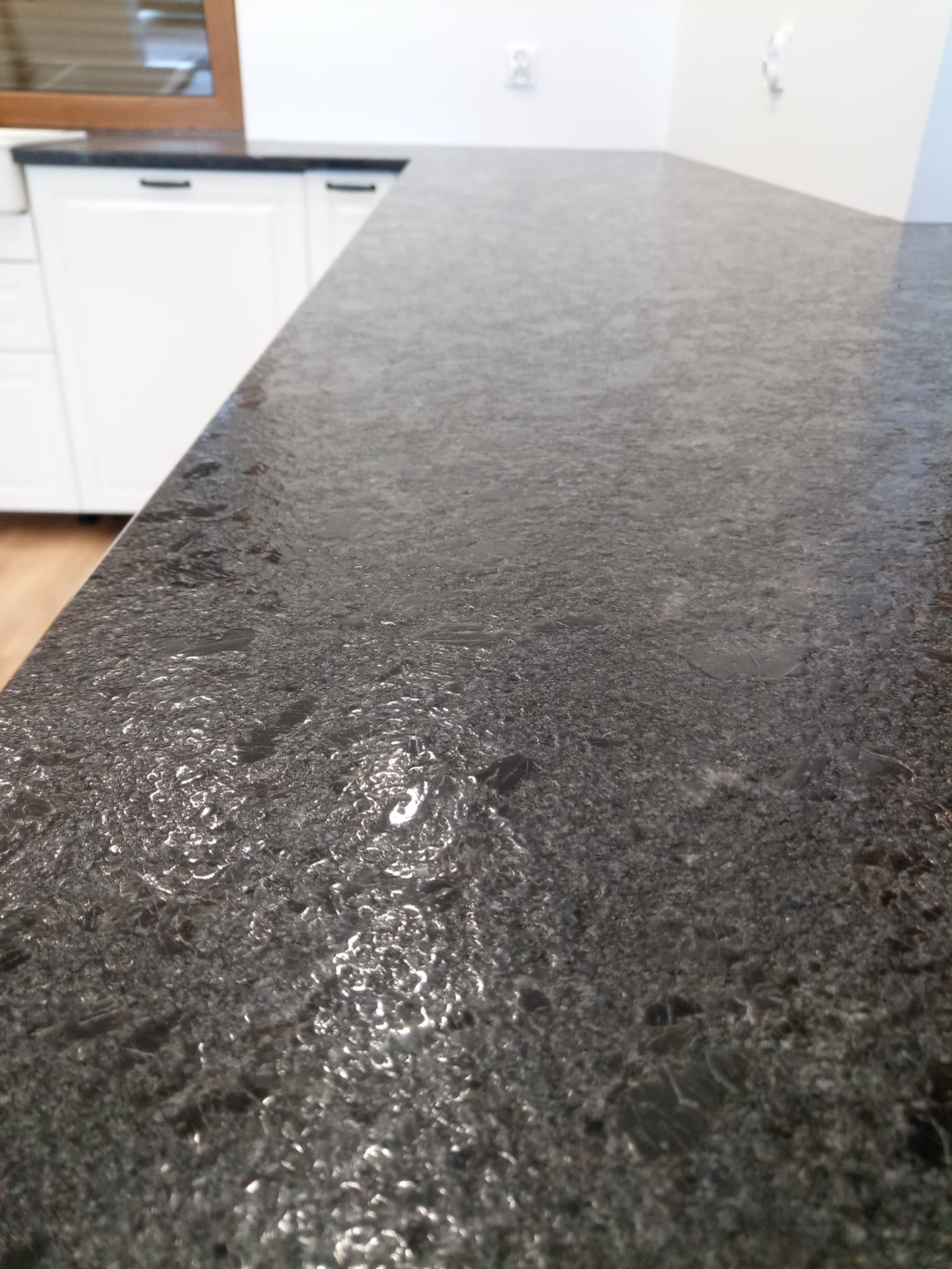 Błyszczący, ciemny blat kamienny z widoczną strukturą, zamontowany w kuchni z białymi szafkami. Ujęcie z bliska, ukazujące detale powierzchni.