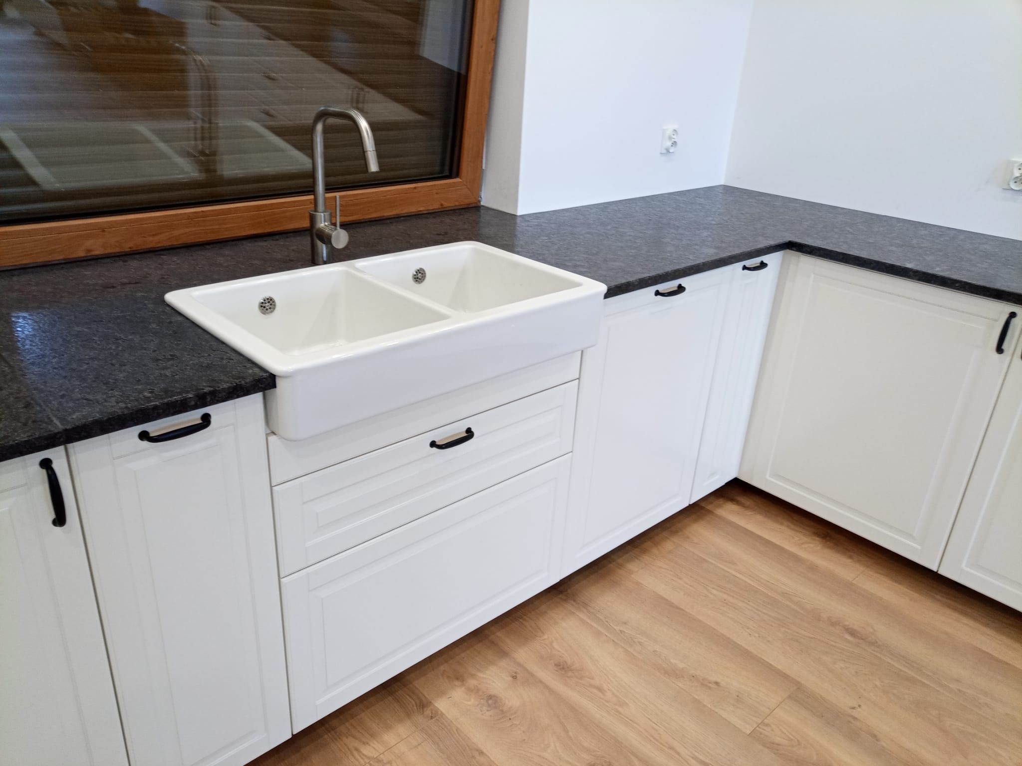 Kuchnia z blatem z ciemnego kamienia, białe szafki z czarnymi uchwytami, ceramiczny zlew dwukomorowy i bateria. Widok na drewniane okno i podłogę.