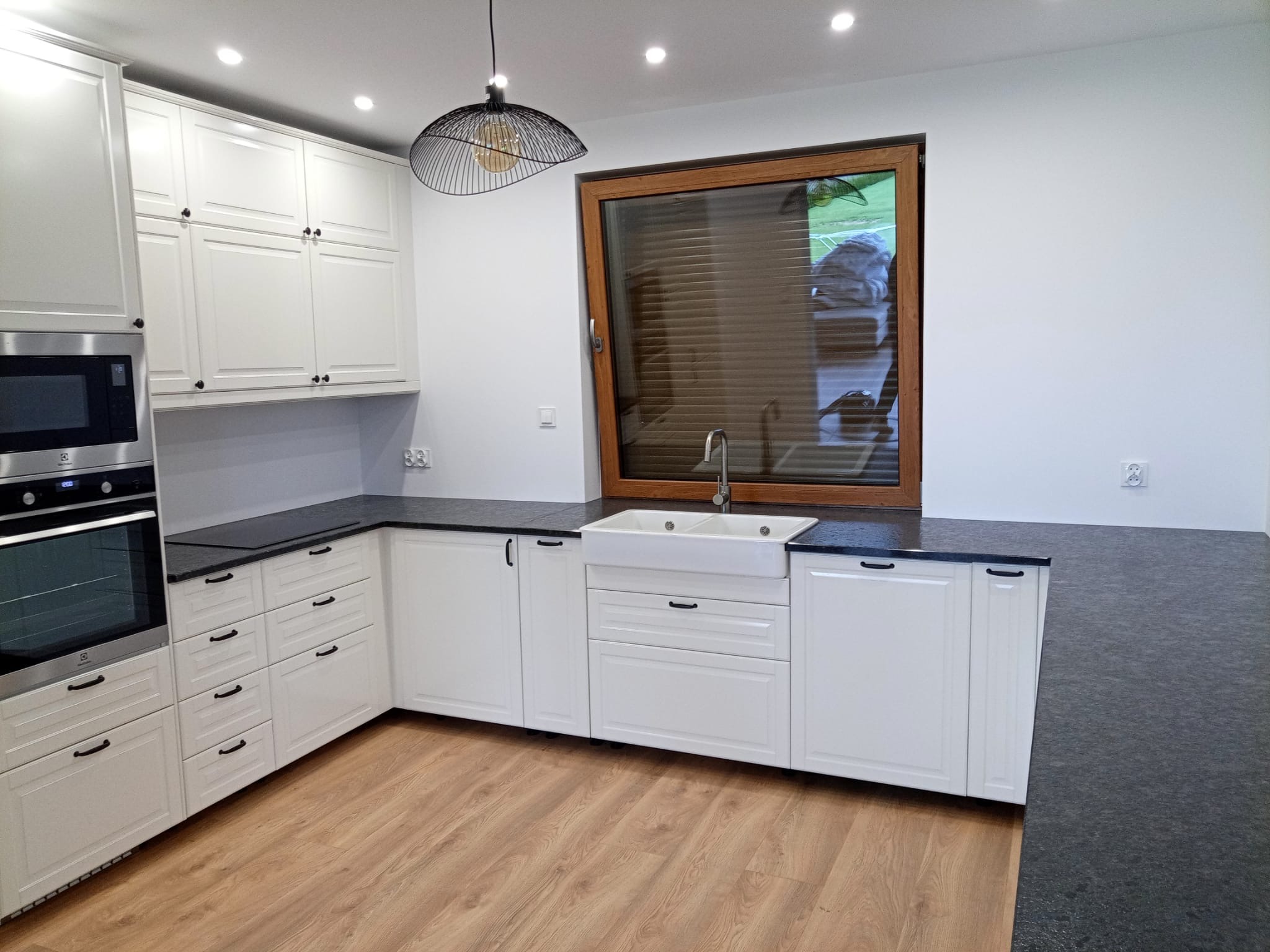 Jasna kuchnia w stylu klasycznym z czarnym blatem, widok na zlew i okno. Uwagę przyciąga designerska lampa wisząca nad zlewem. Podłoga drewniana.