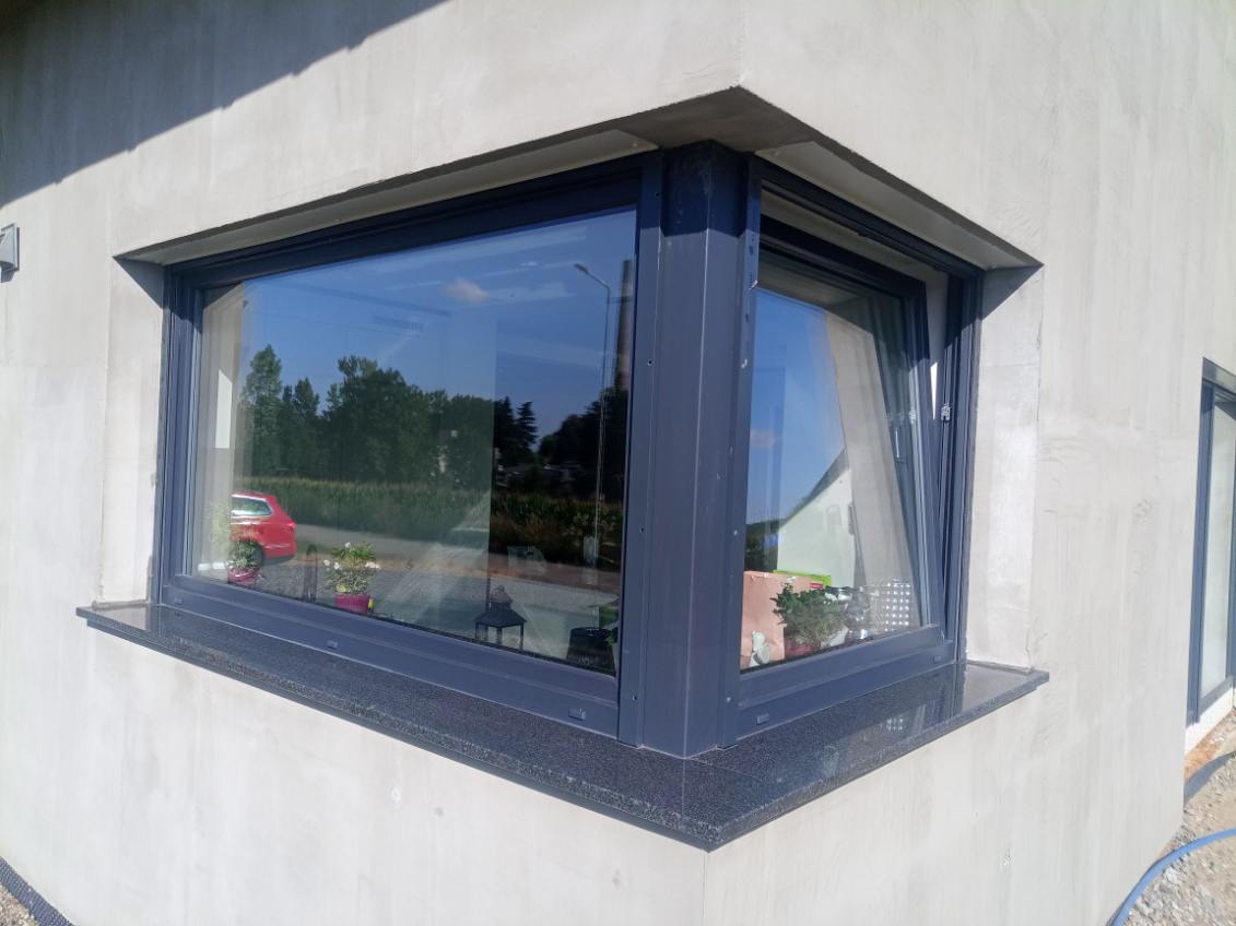 Granitowy parapet zewnętrzny, ciemny, ziarnisty, pod oknem narożnym w surowym budynku. Widok z zewnątrz, w tle zieleń i odbicie nieba w szybie.