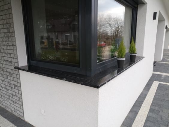 Czarny parapet zewnętrzny z kamienia, zdobiący okno budynku z szarą elewacją. Na parapecie ustawione dwie małe, zielone rośliny w doniczkach.