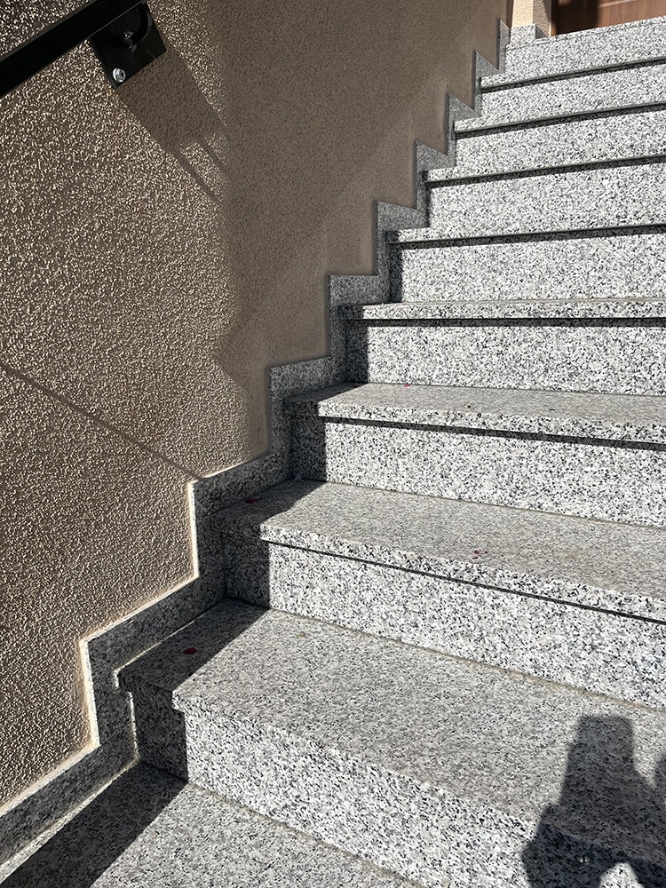 Zewnętrzne schody z jasnego, nakrapianego kamienia prowadzą w górę, przylegając do ściany o szorstkiej fakturze. Metalowa poręcz widoczna w lewym górnym rogu.