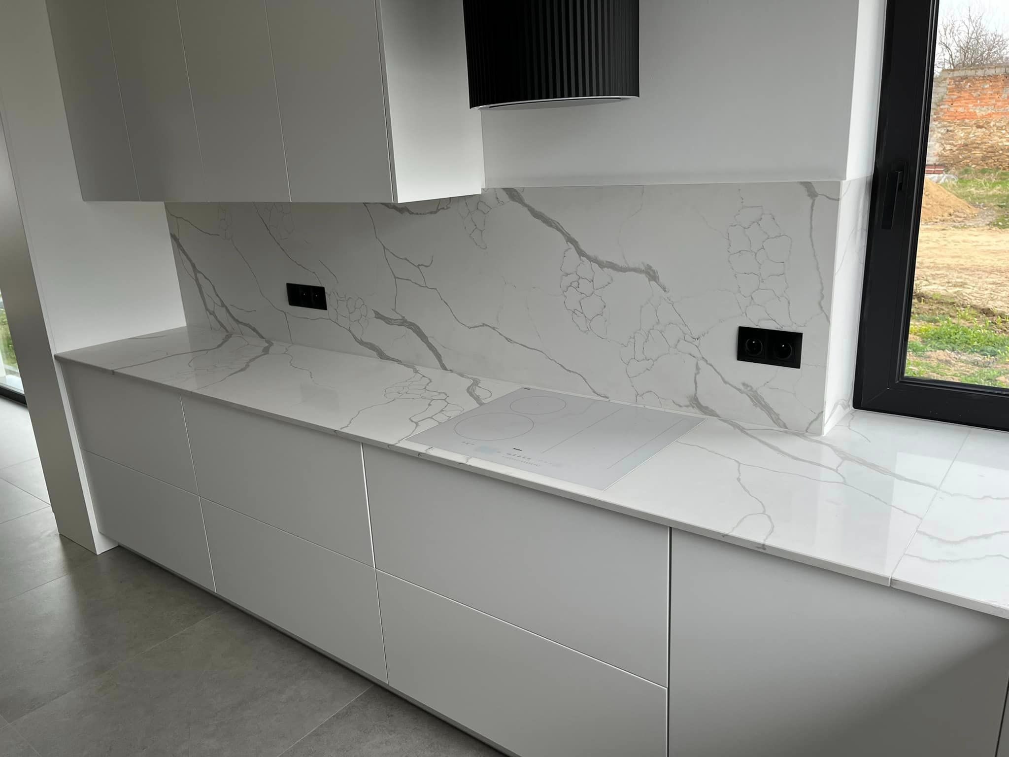 Biały blat kamienny z marmurowym wzorem w nowoczesnej kuchni. Widoczna płyta indukcyjna i okap. Minimalistyczny design, jasne wnętrze z widokiem na zewnątrz.