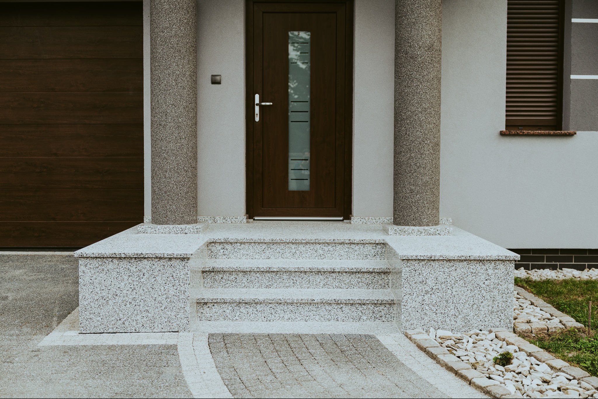 Kamienne schody wejściowe z jasnego granitu prowadzące do brązowych drzwi z przeszkleniem. Kolumny z tego samego kamienia po bokach. Nowoczesny design.