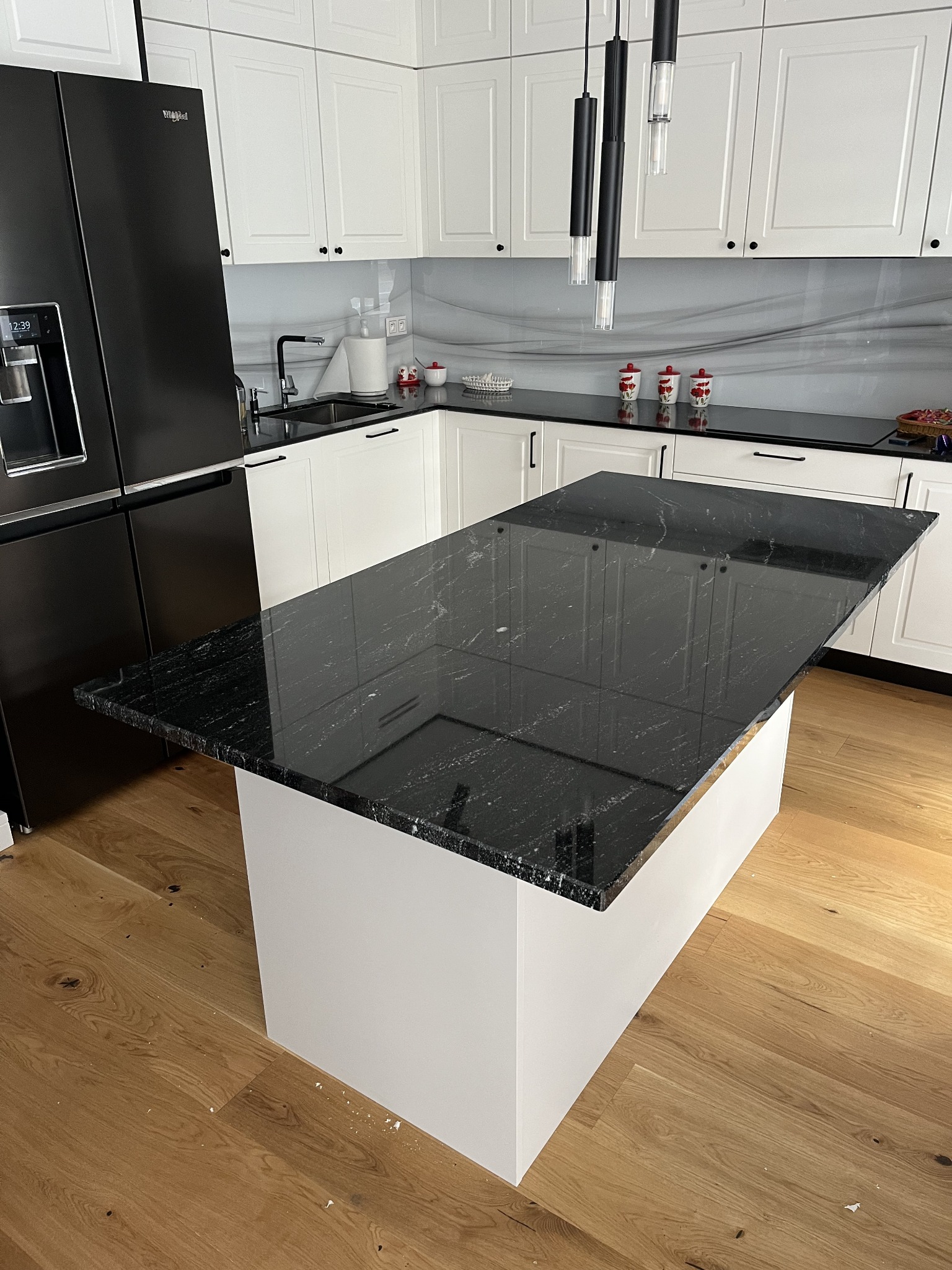 Czarny, błyszczący blat kamienny na wyspie kuchennej z białym korpusem, w nowoczesnej kuchni z drewnianą podłogą i białymi szafkami. Widoczne lampy wiszące.