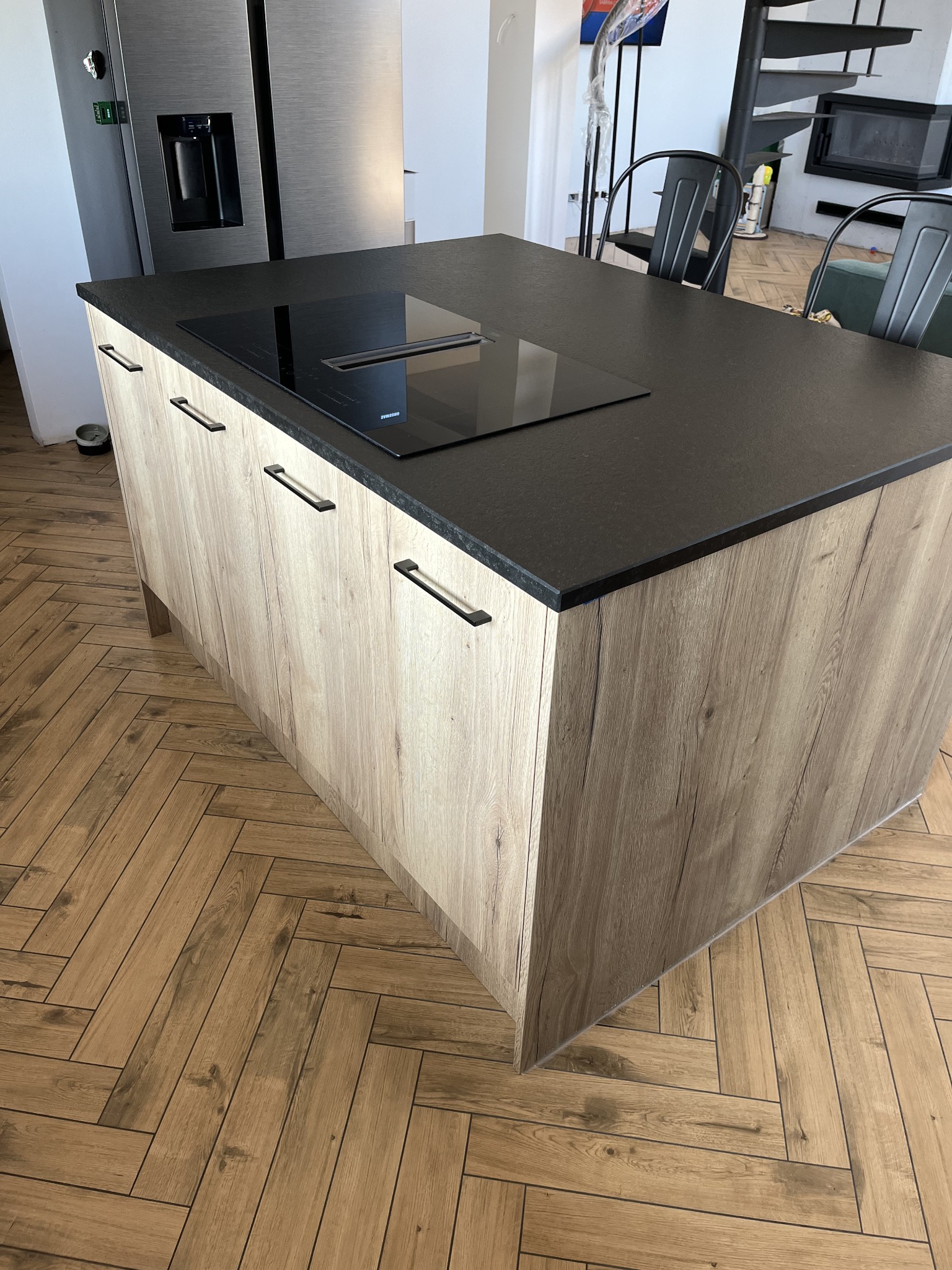 Wyspa kuchenna z blatem z ciemnego kamienia i drewnopodobnymi frontami szafek, na tle parkietu ułożonego w jodełkę. Widoczna płyta indukcyjna i fragment spiralnych schodów.